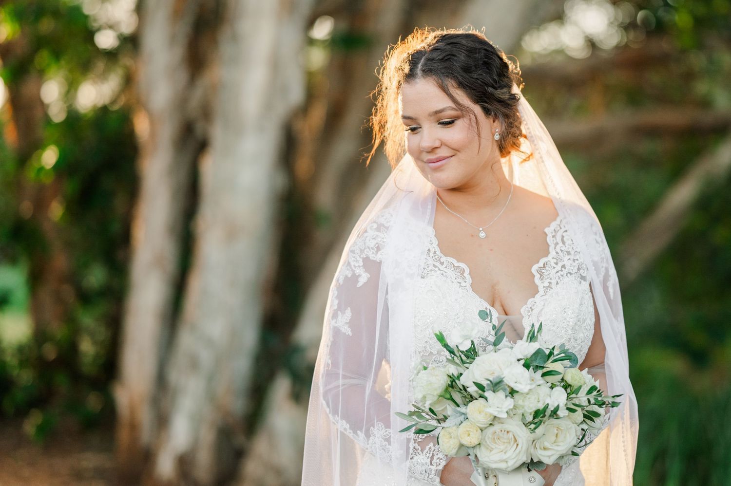 A bridal portrait showcasing a white lace wedding gown and delicate bouquet against a sunlit natural backdrop.