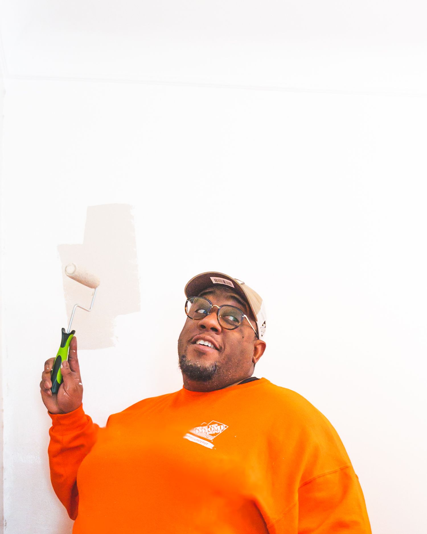 Person in bright orange Home Depot sweatshirt holding painting supplies against white wall.