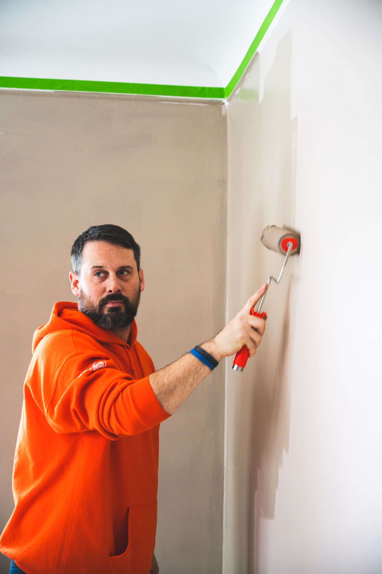 Person in orange hoodie using paint roller on interior wall with green painter's tape.