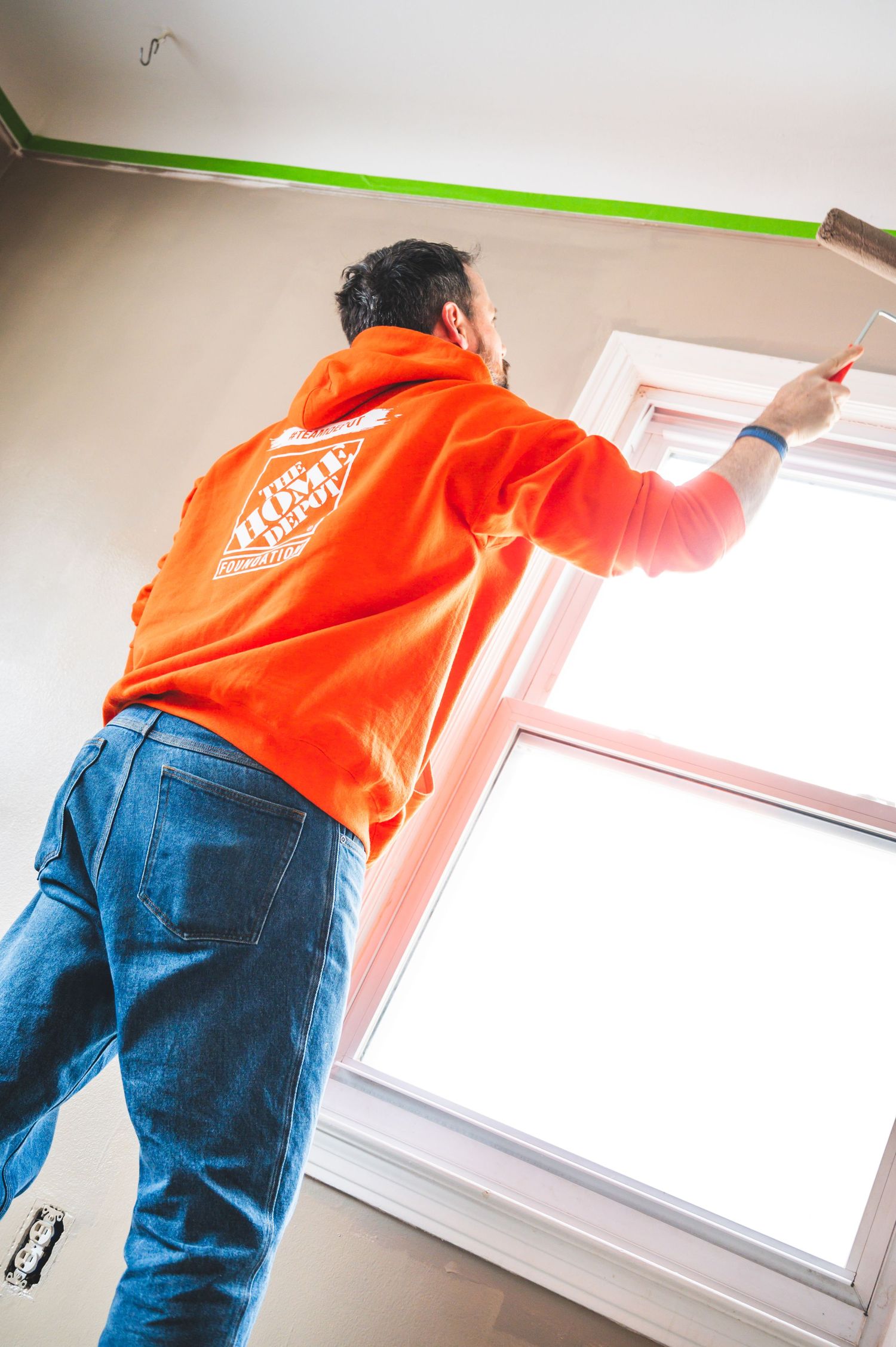 Person in orange Home Depot shirt painting near a window with green tape for trim work.