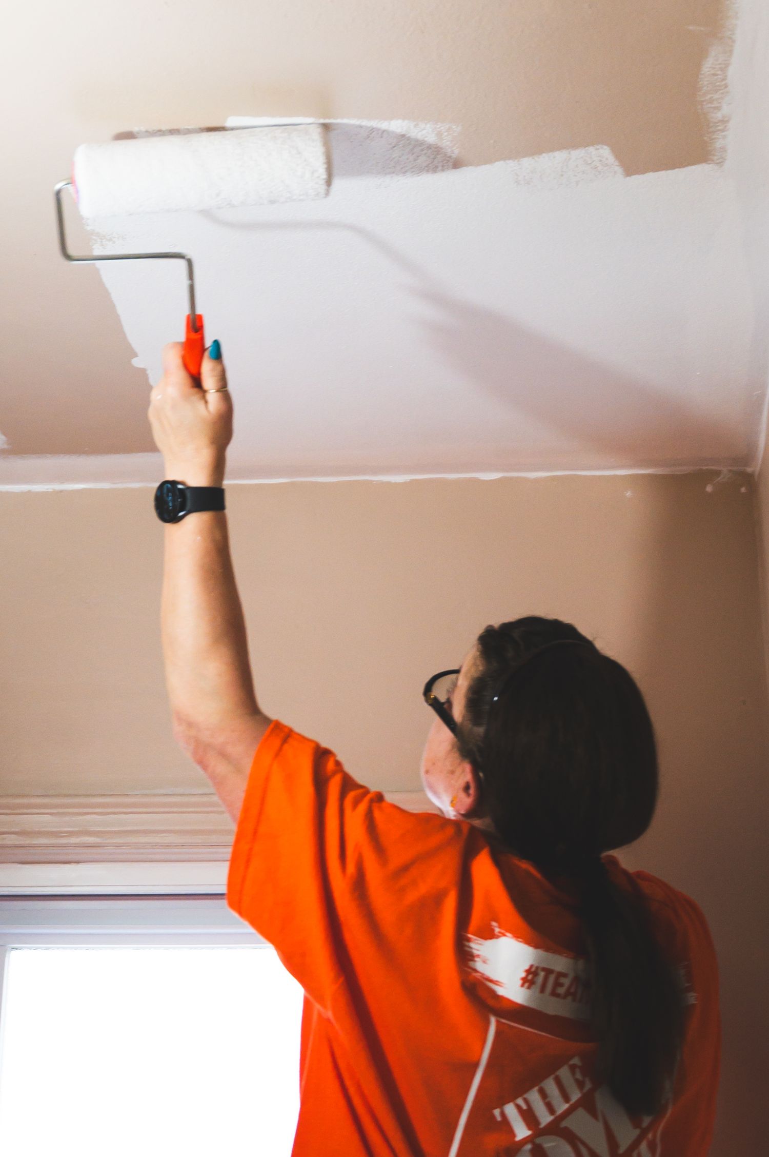 Person in orange t-shirt using paint roller to apply white paint to ceiling.