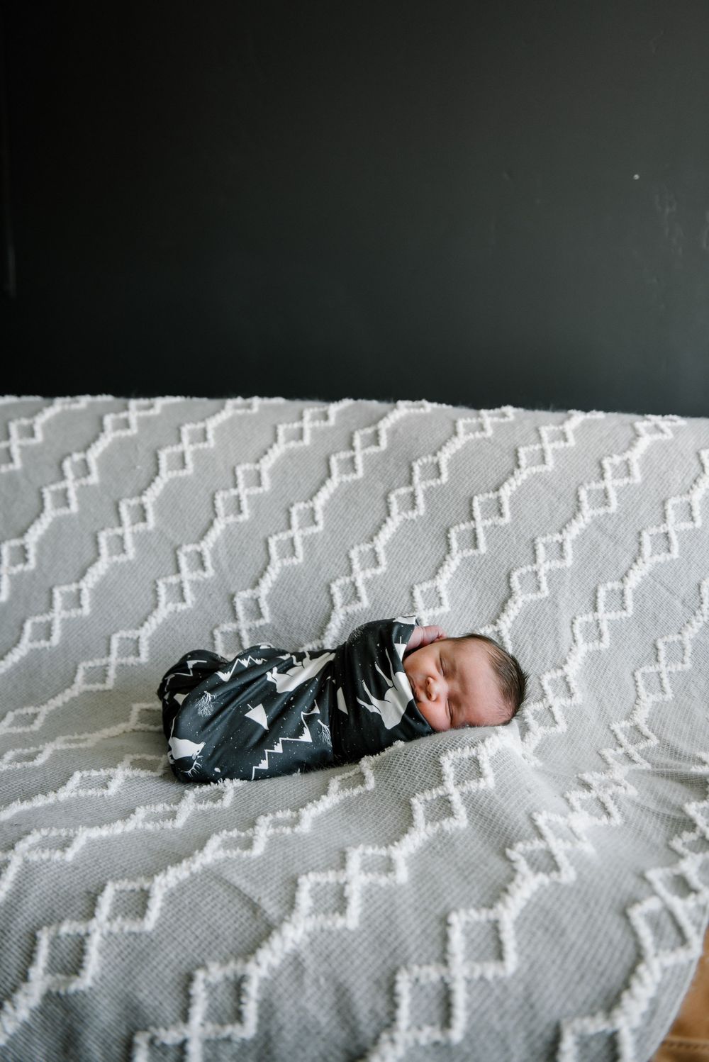 Swaddled newborn resting on white geometric patterned blanket.