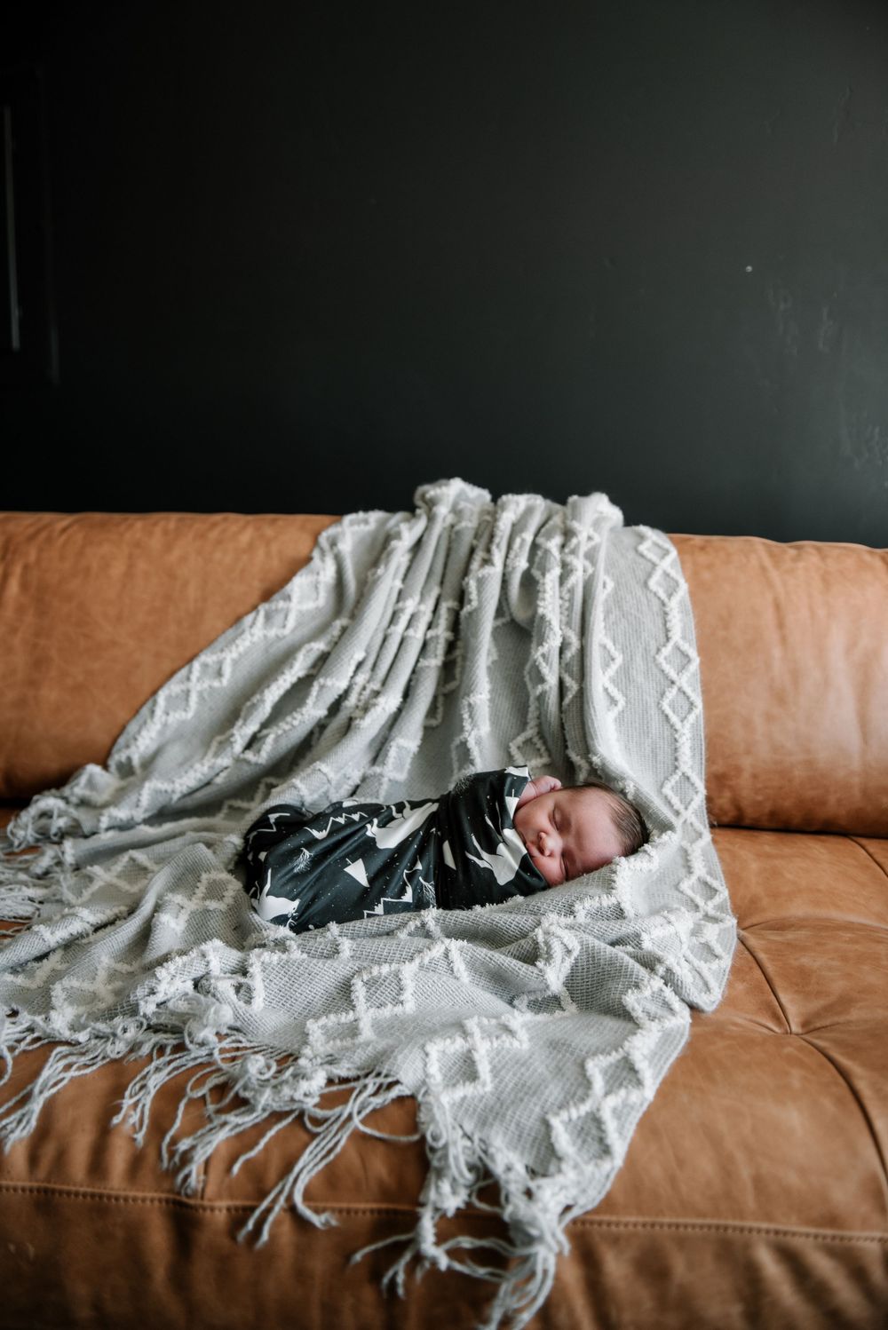 Sleeping baby wrapped in black swaddle on brown leather couch with textured blanket.
