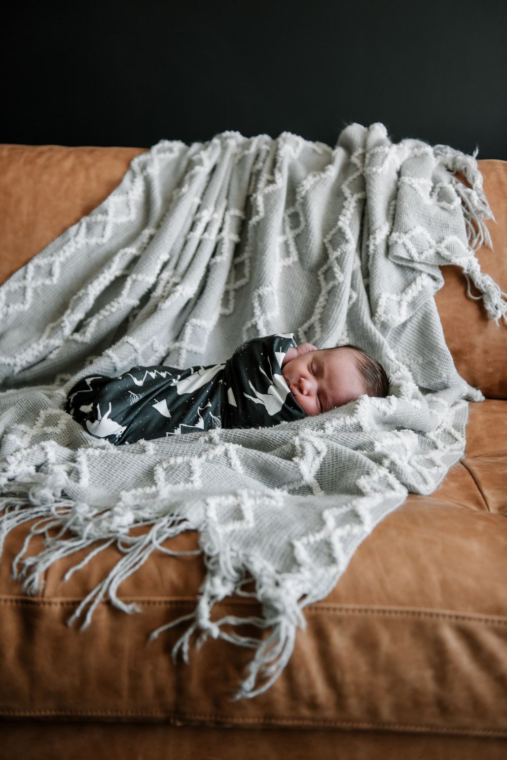 Newborn sleeping peacefully on brown couch covered with patterned gray throw blanket.