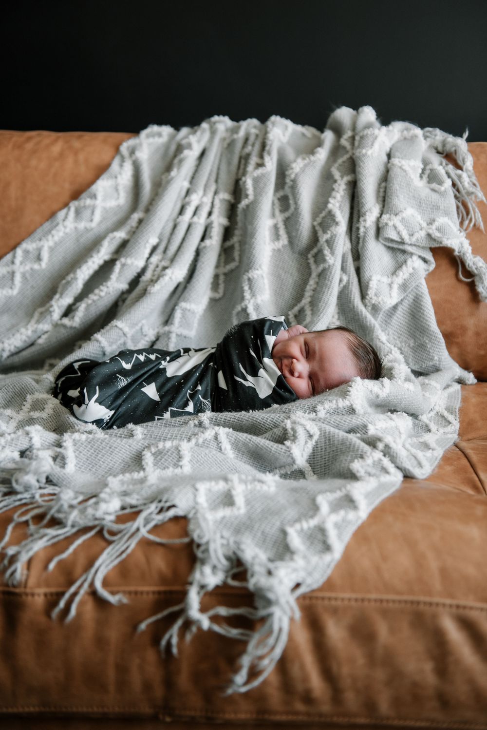 Sleeping baby wrapped in white knit blanket resting on brown leather couch.
