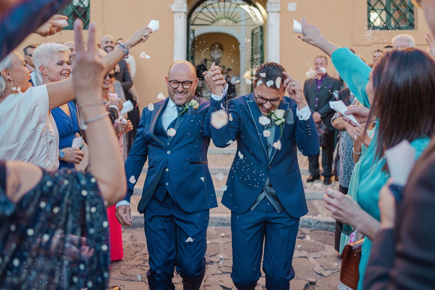 Wedding guests shower a celebrating couple with confetti outside a grand building.