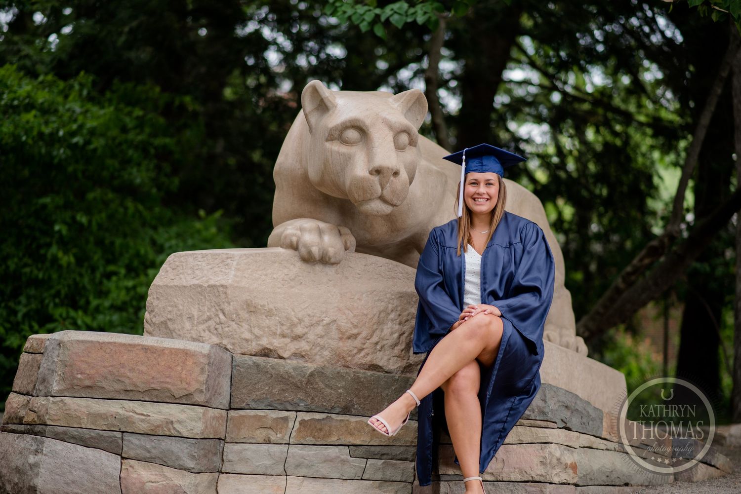 Graduate in blue regalia sits on stone ledge next to iconic Penn State lion statue.