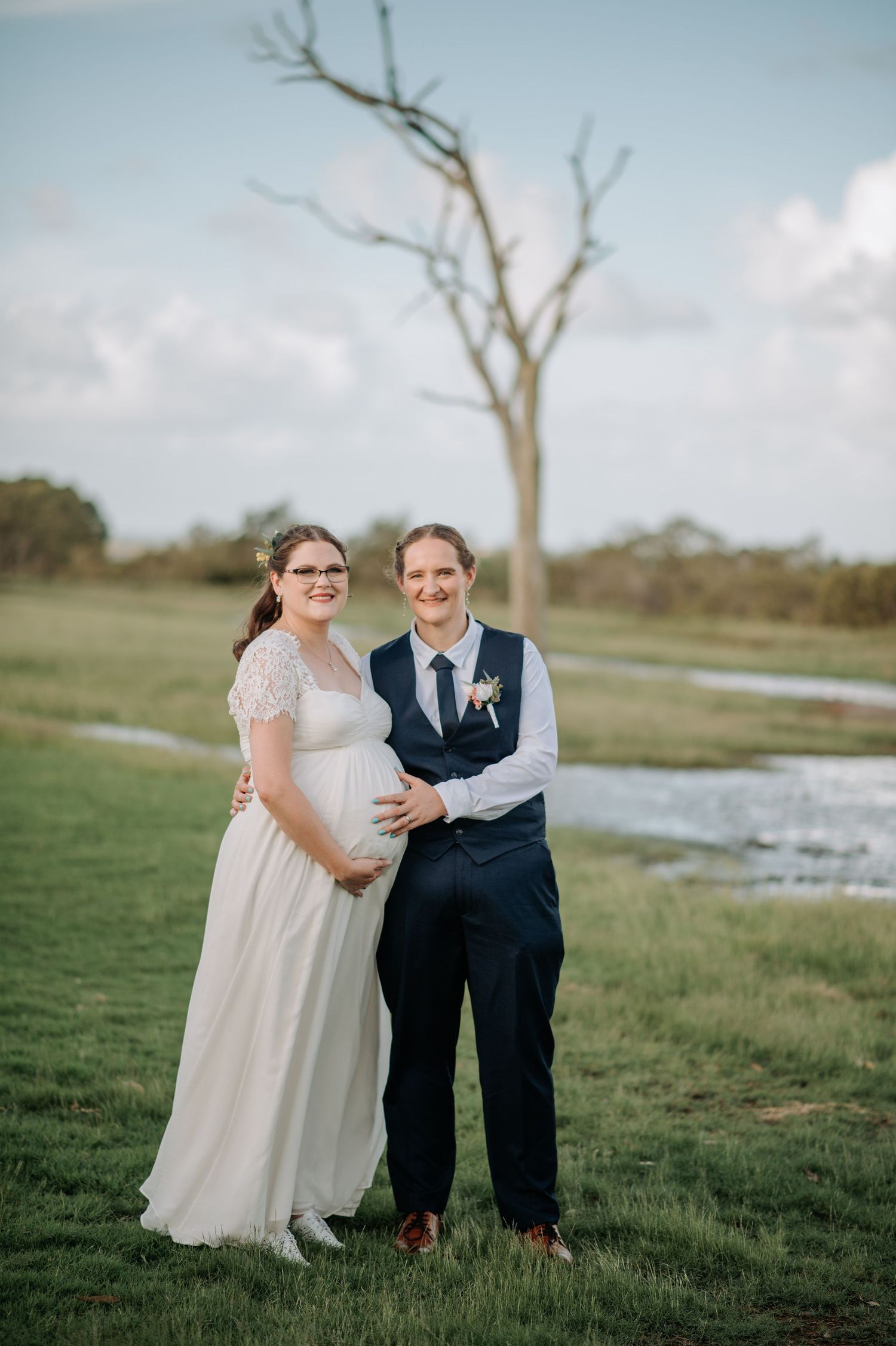 Couple poses together in scenic field with bare tree in background.