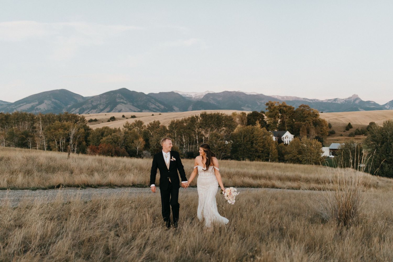 A countryside wedding portrait showing a couple walking through tall grass with mountains behind.