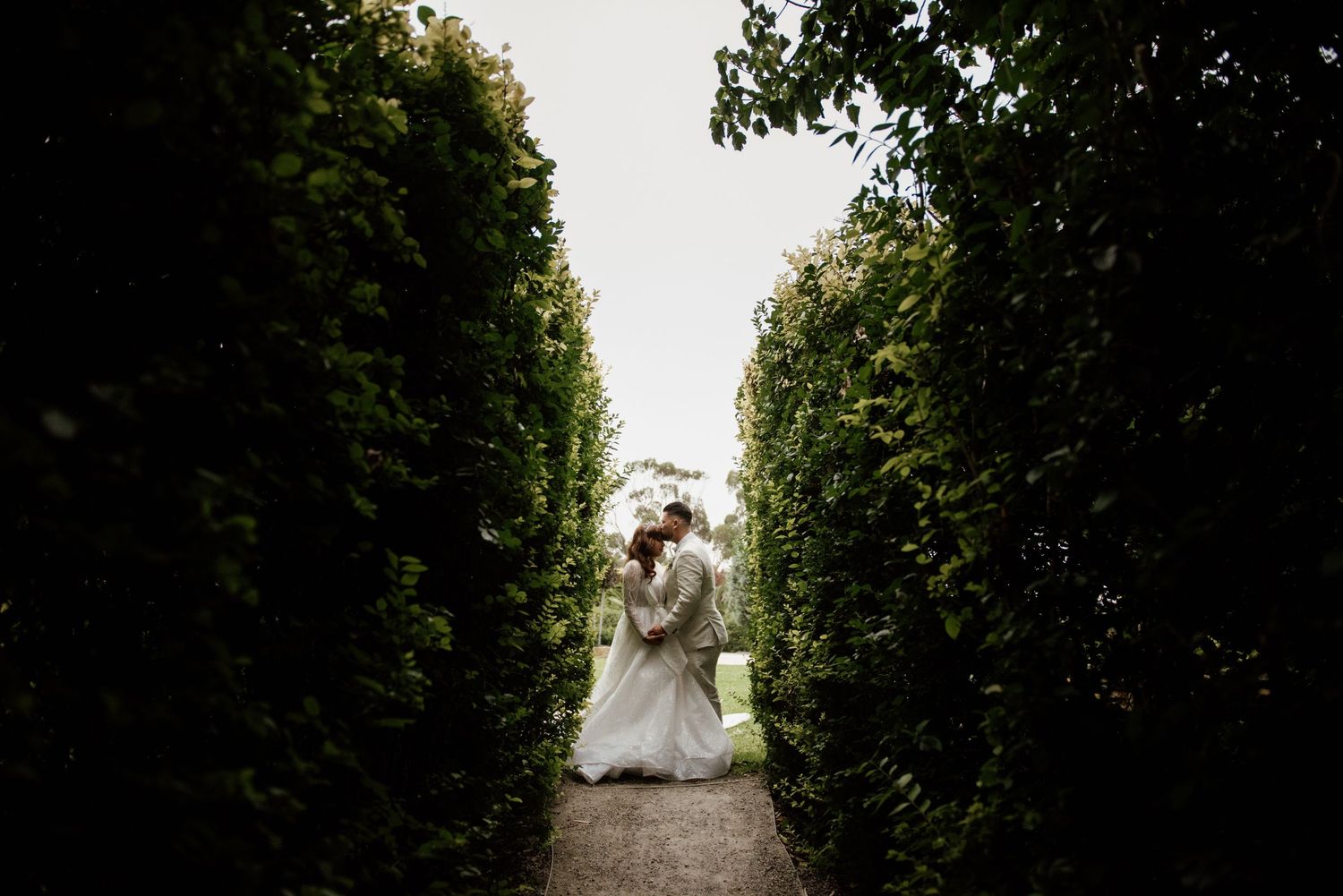 A couple embraces in a garden path framed by tall manicured hedges.
