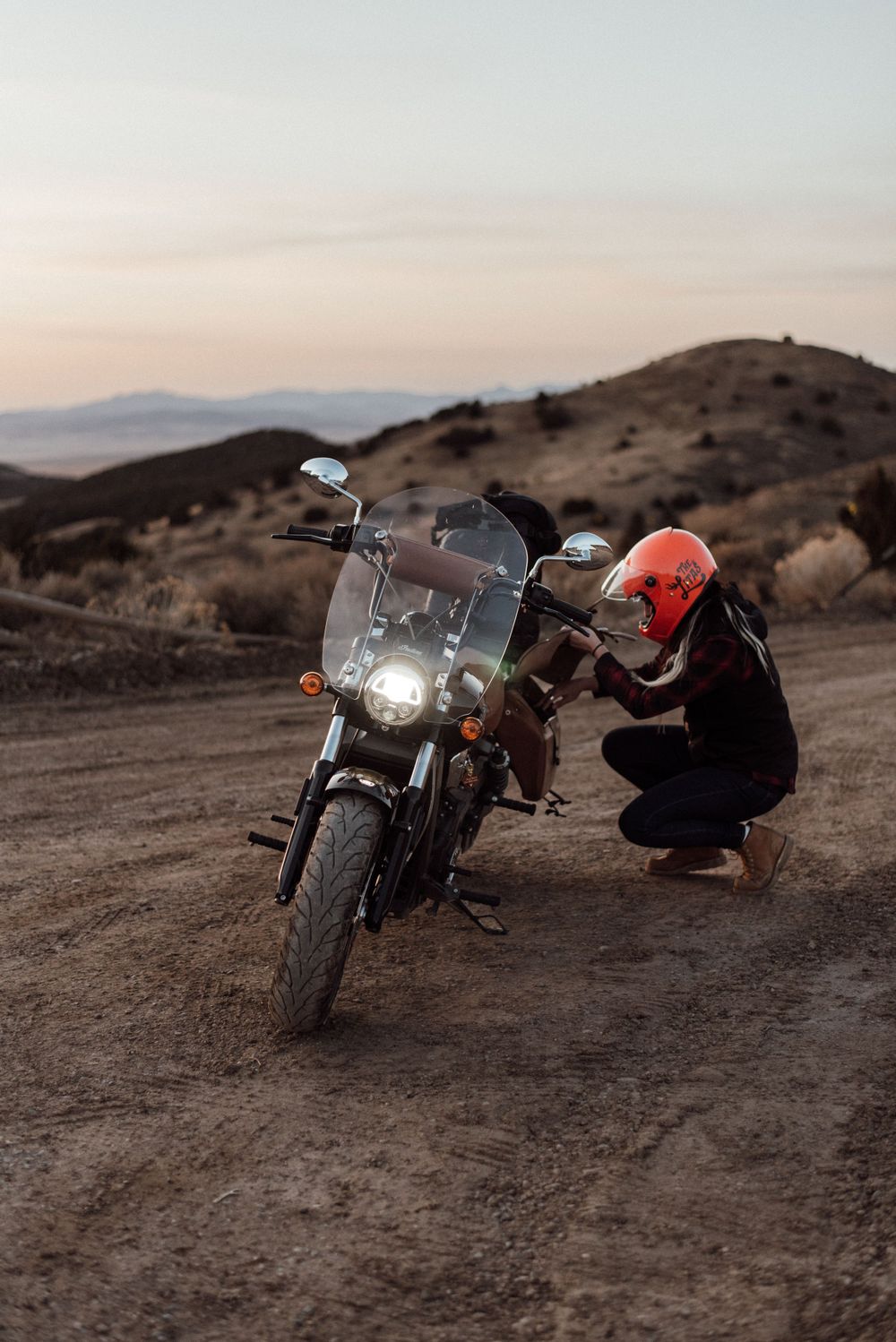 Motorcyclist in red helmet inspecting a parked motorcycle on a dirt trail at sunset with mountains in background.