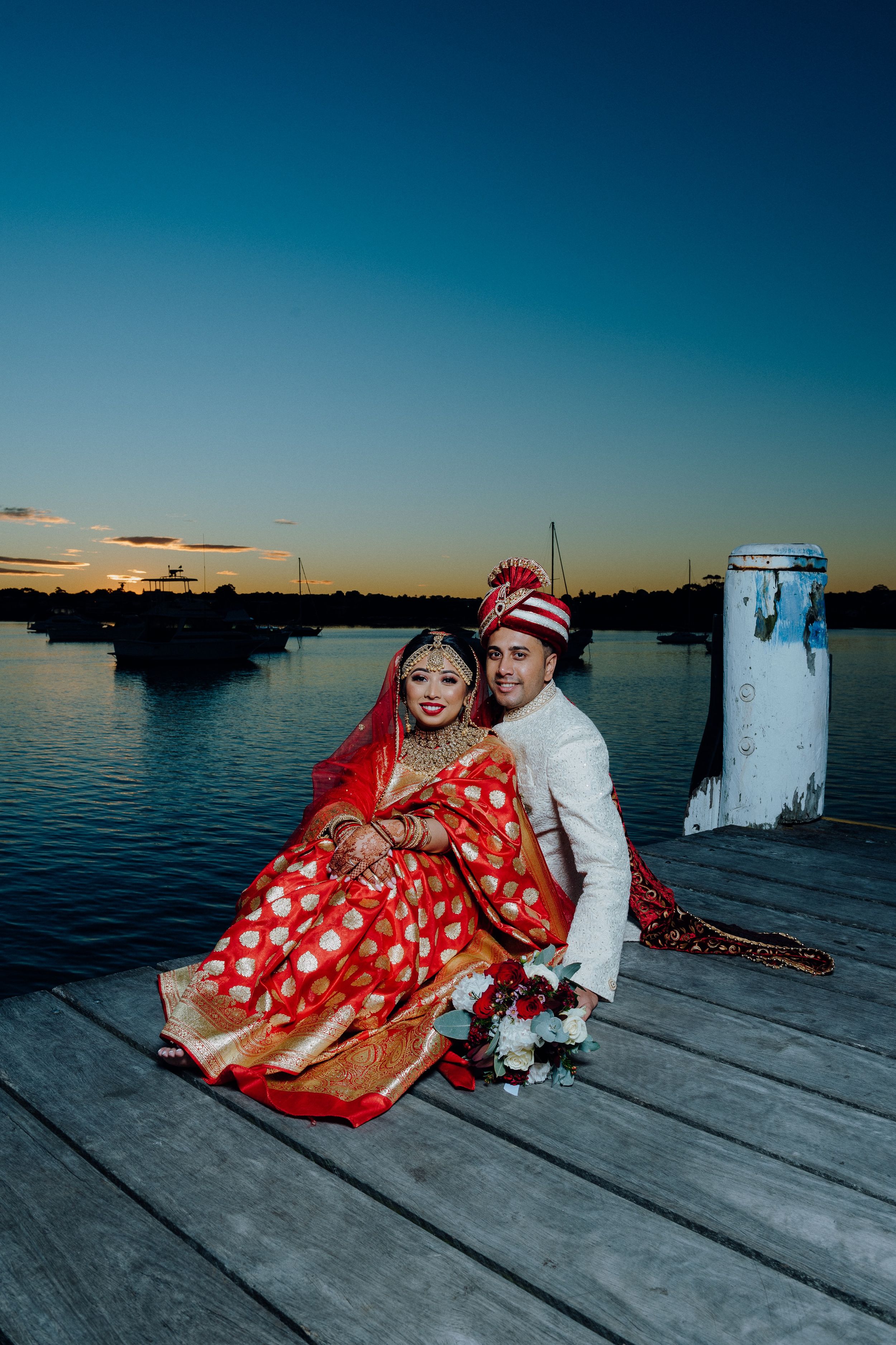 A couple in traditional South Asian wedding attire sitting on a wooden dock at sunset.