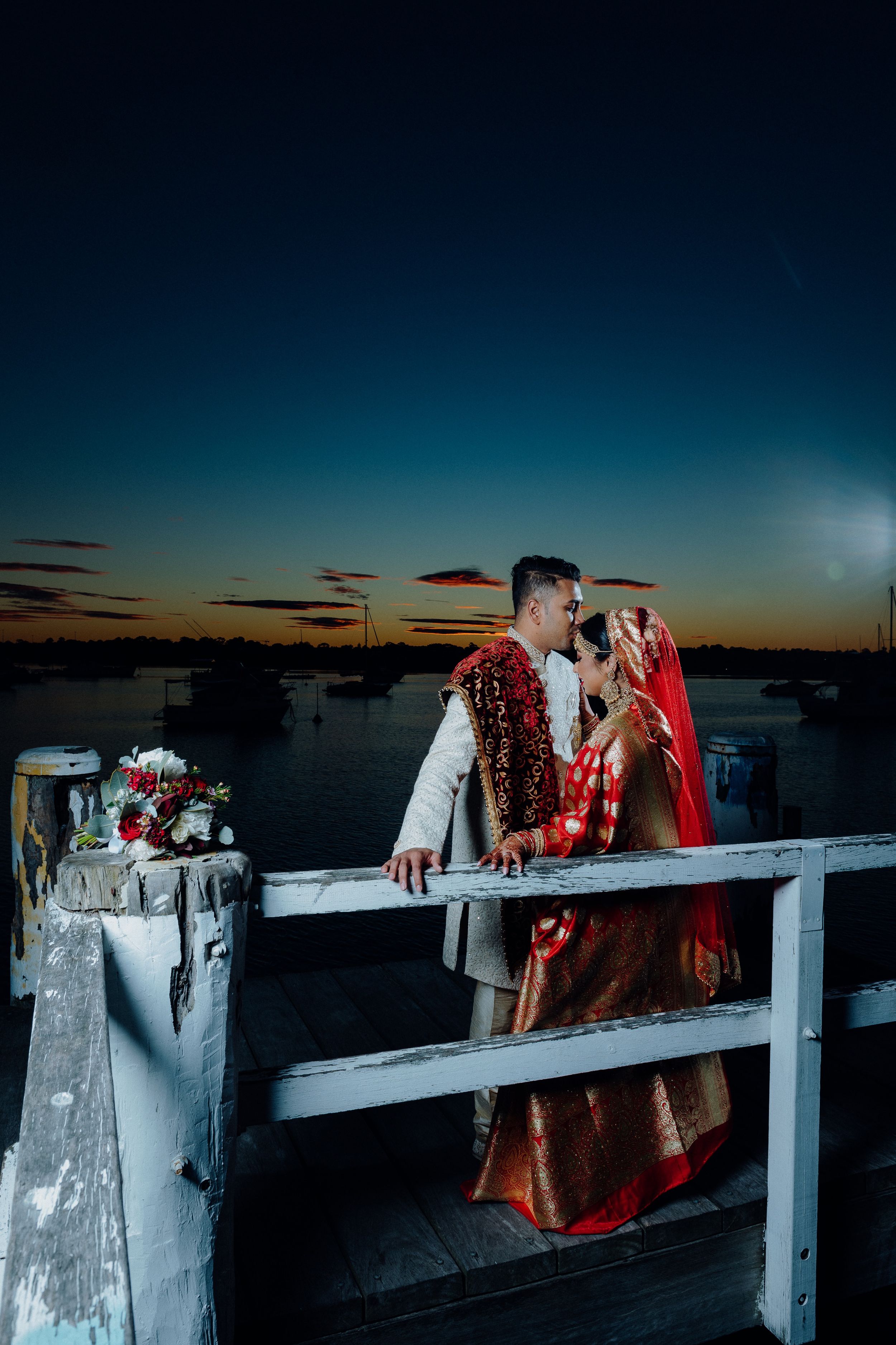 Couple in traditional wedding attire pose romantically on dock during sunset.
