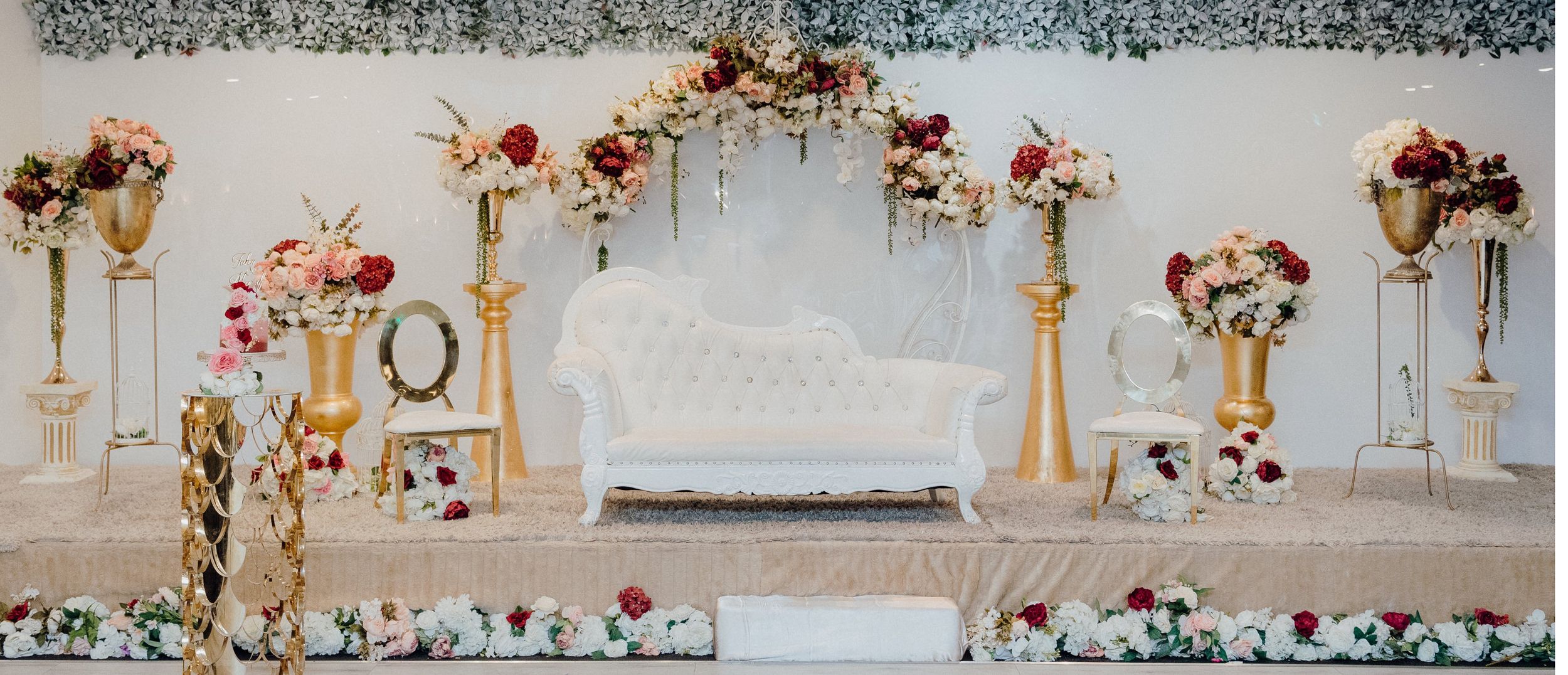 Elegant wedding stage setup with white sofa, gold pillars, and pink and red floral arrangements.