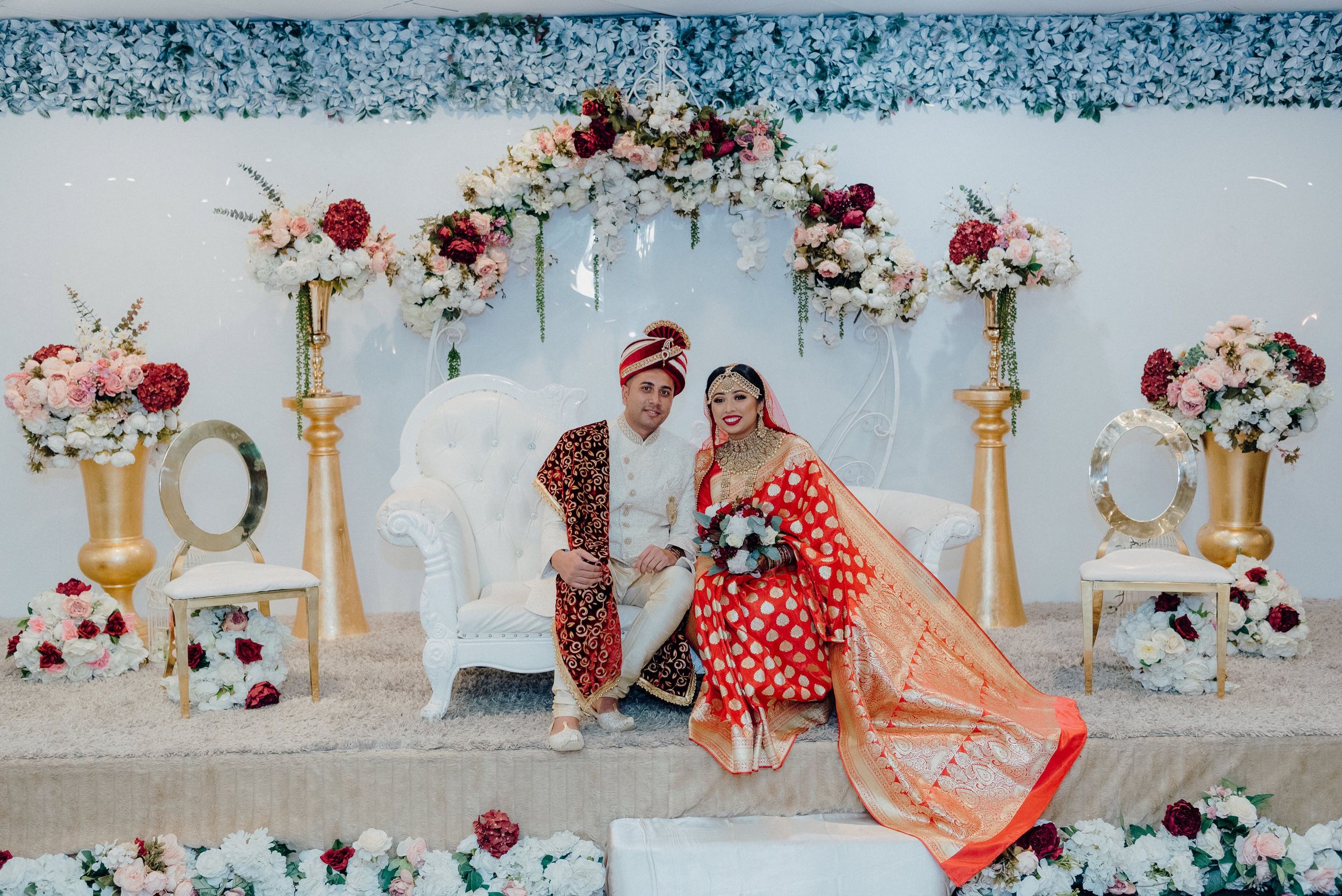 Wedding portrait against white backdrop with gold pillars and pink and red floral arrangements.