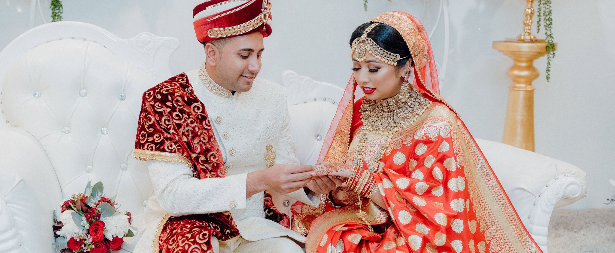 A couple in traditional red and white wedding attire exchanging rings during ceremony.