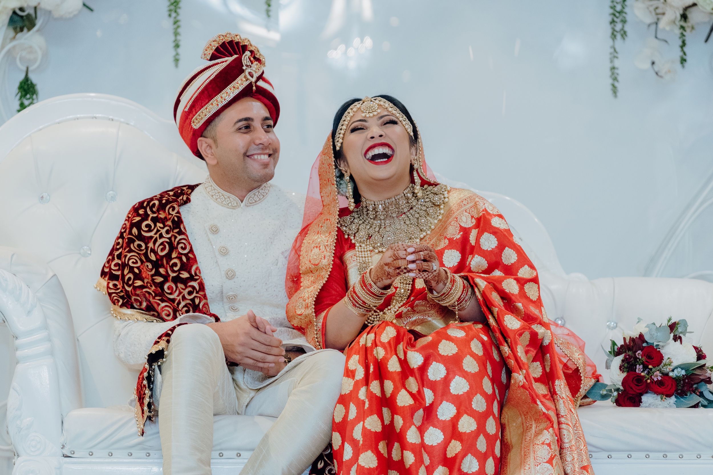 Wedding couple in traditional red and white attire sharing joyful moment on decorated stage.