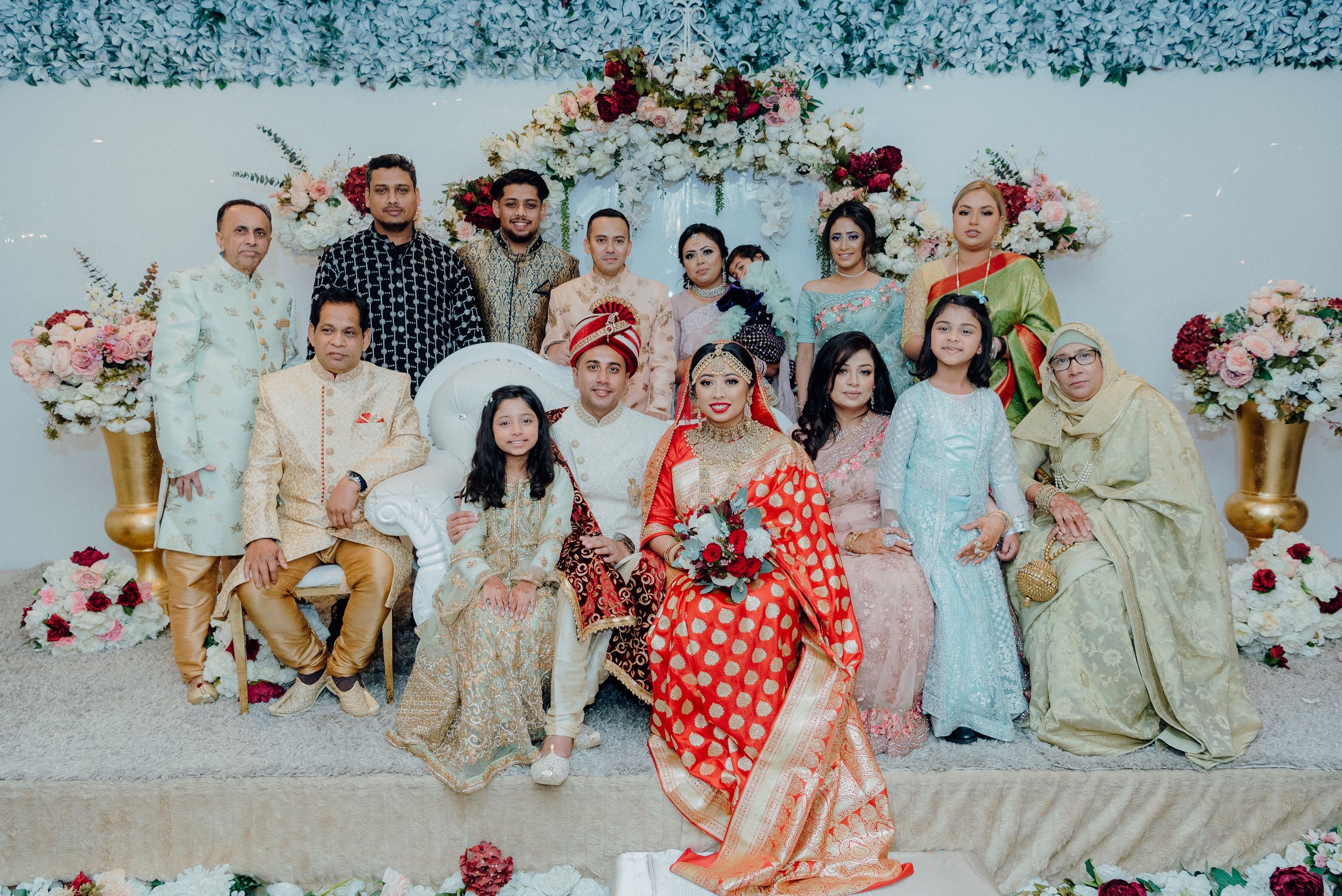 Wedding celebration group portrait with guests in traditional attire against decorated backdrop.
