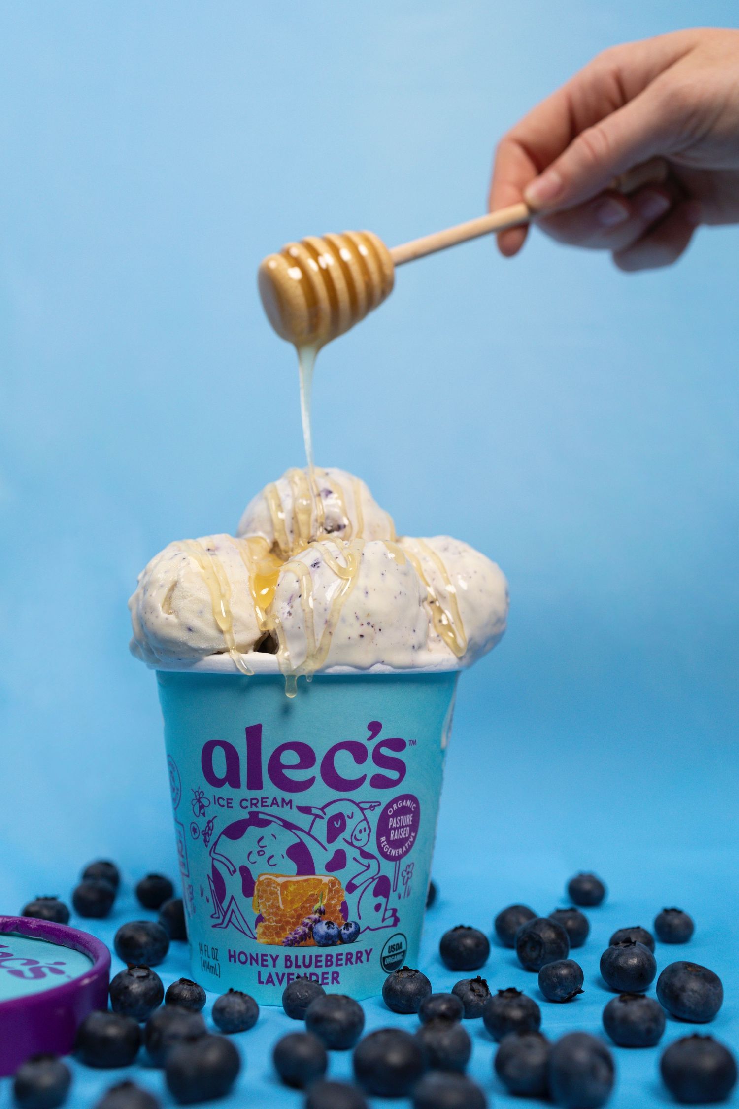 Honey drizzles from a wooden dipper onto Alec's Blueberry Lavender ice cream pint with blueberries.