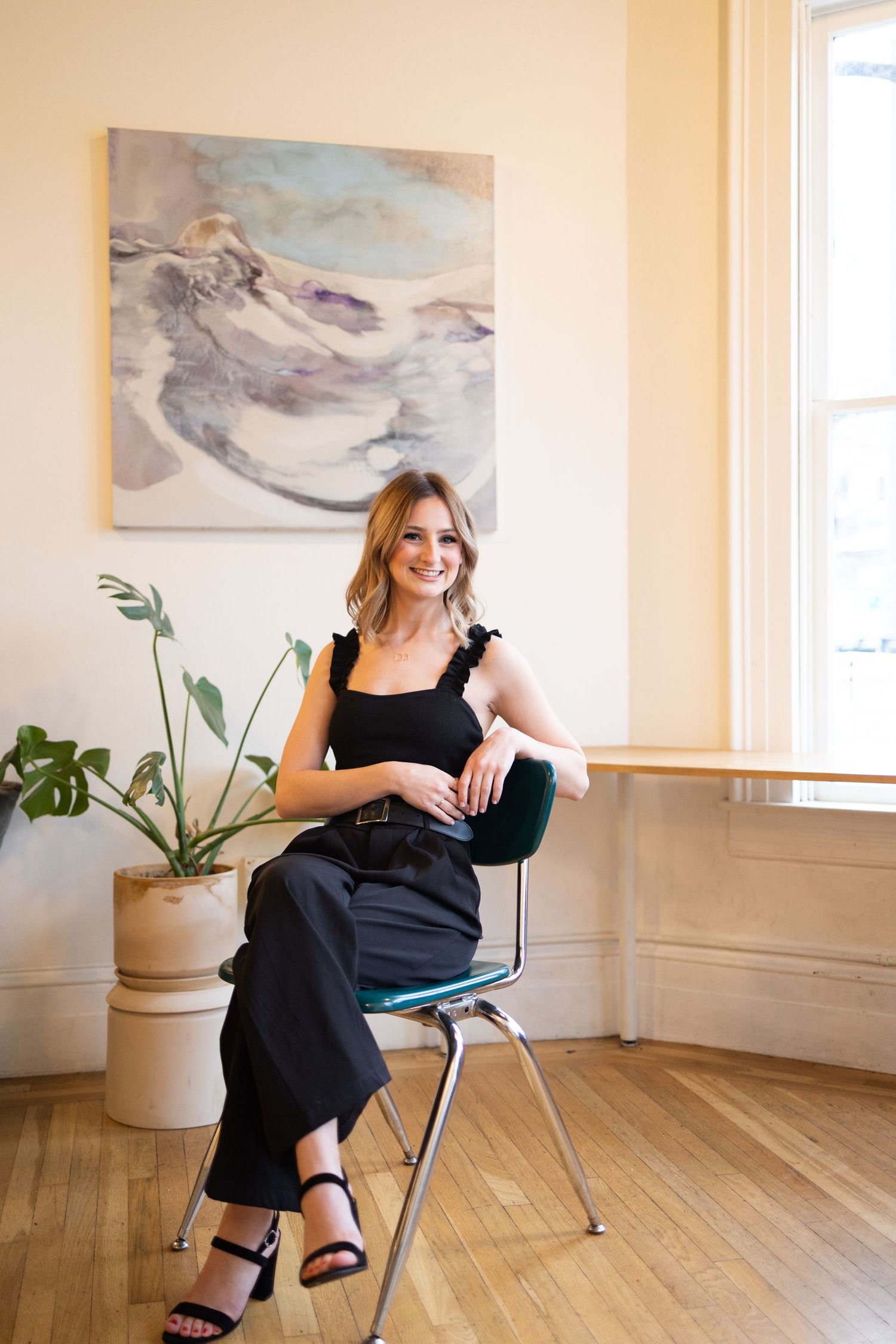 Young woman in black outfit sitting on chair in bright room with abstract wall art.
