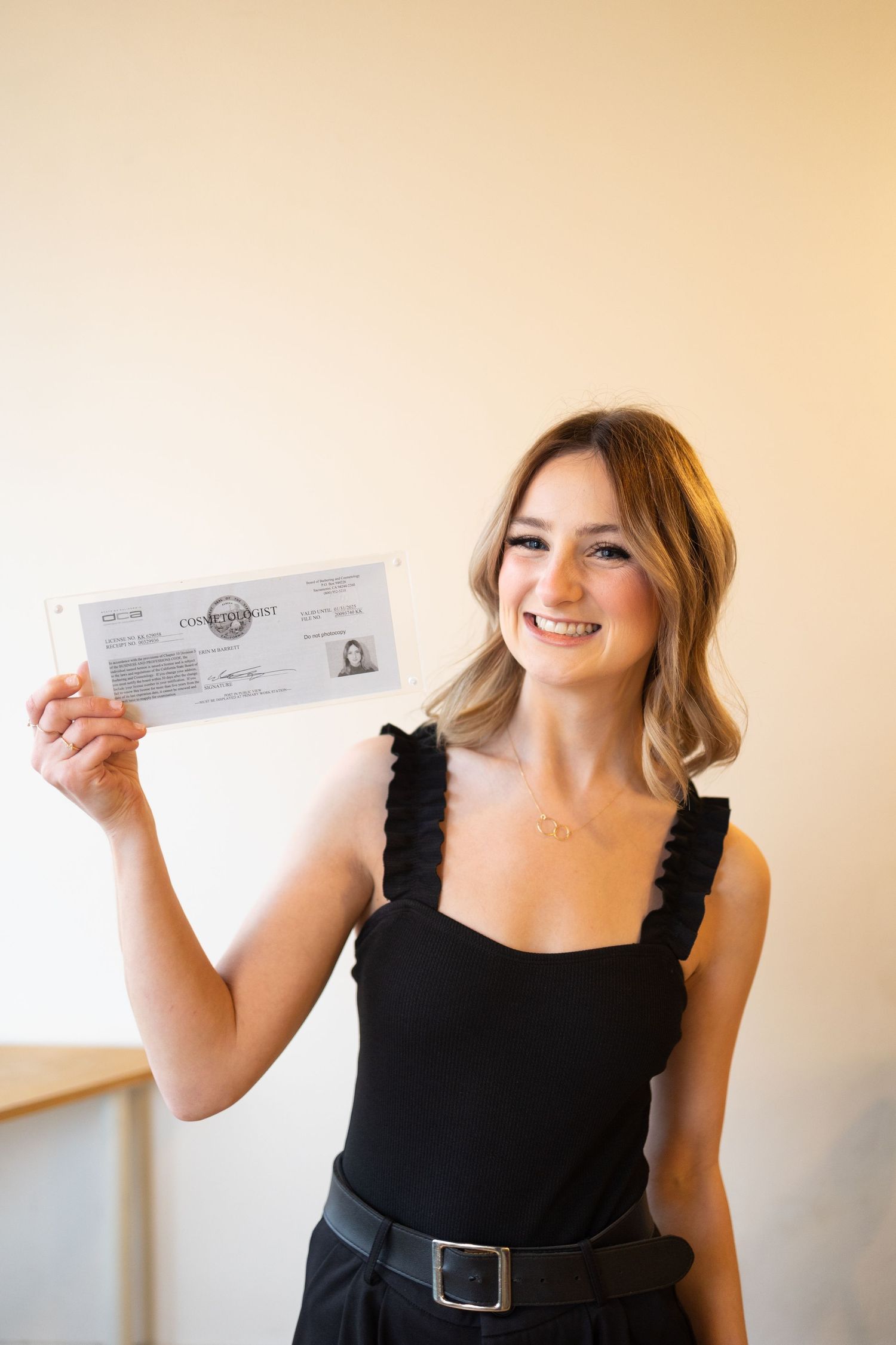 Smiling blonde woman in black top holding up a document or certificate indoors.