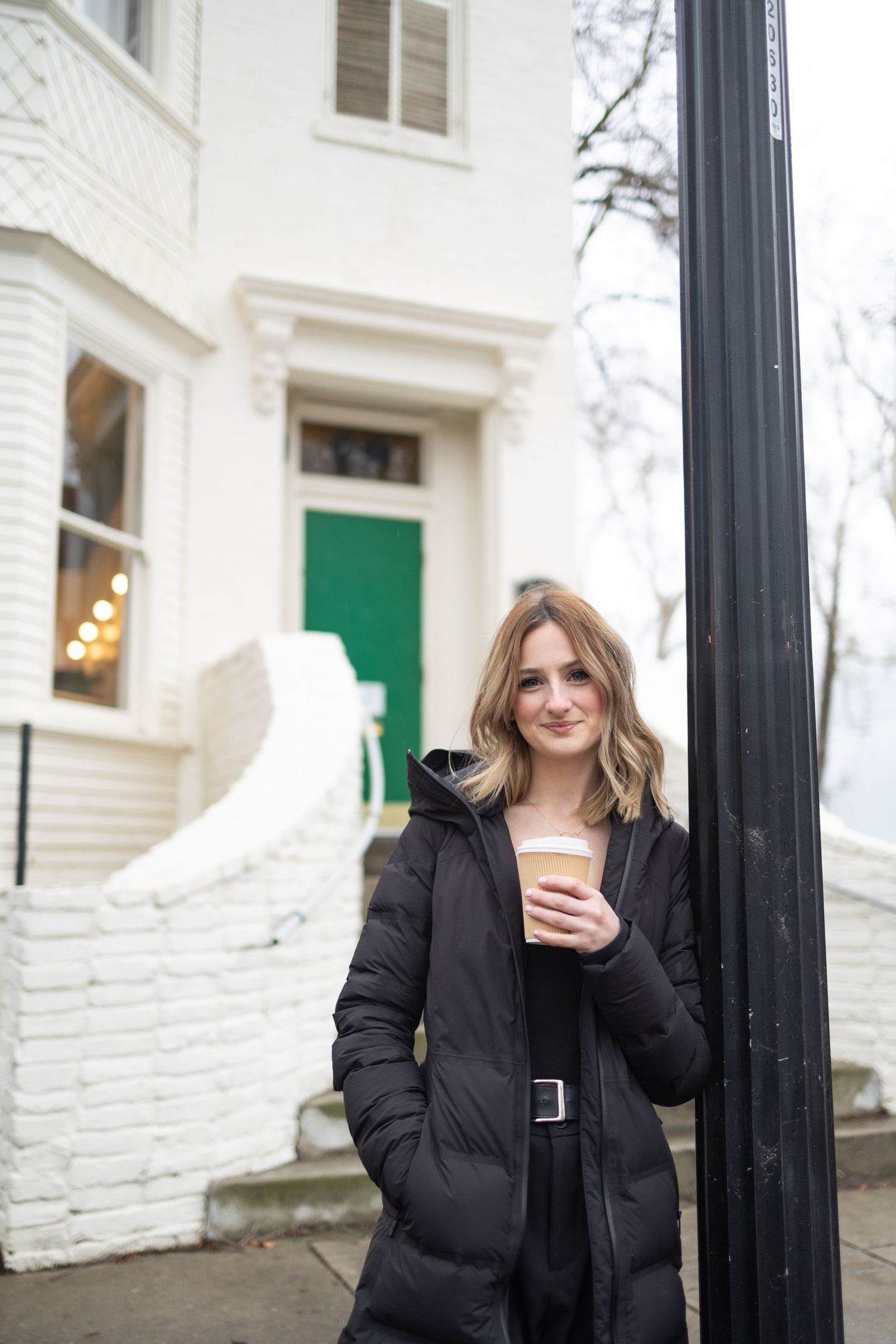 Blonde woman in black puffer coat holding coffee cup leaning on lamppost on city street.