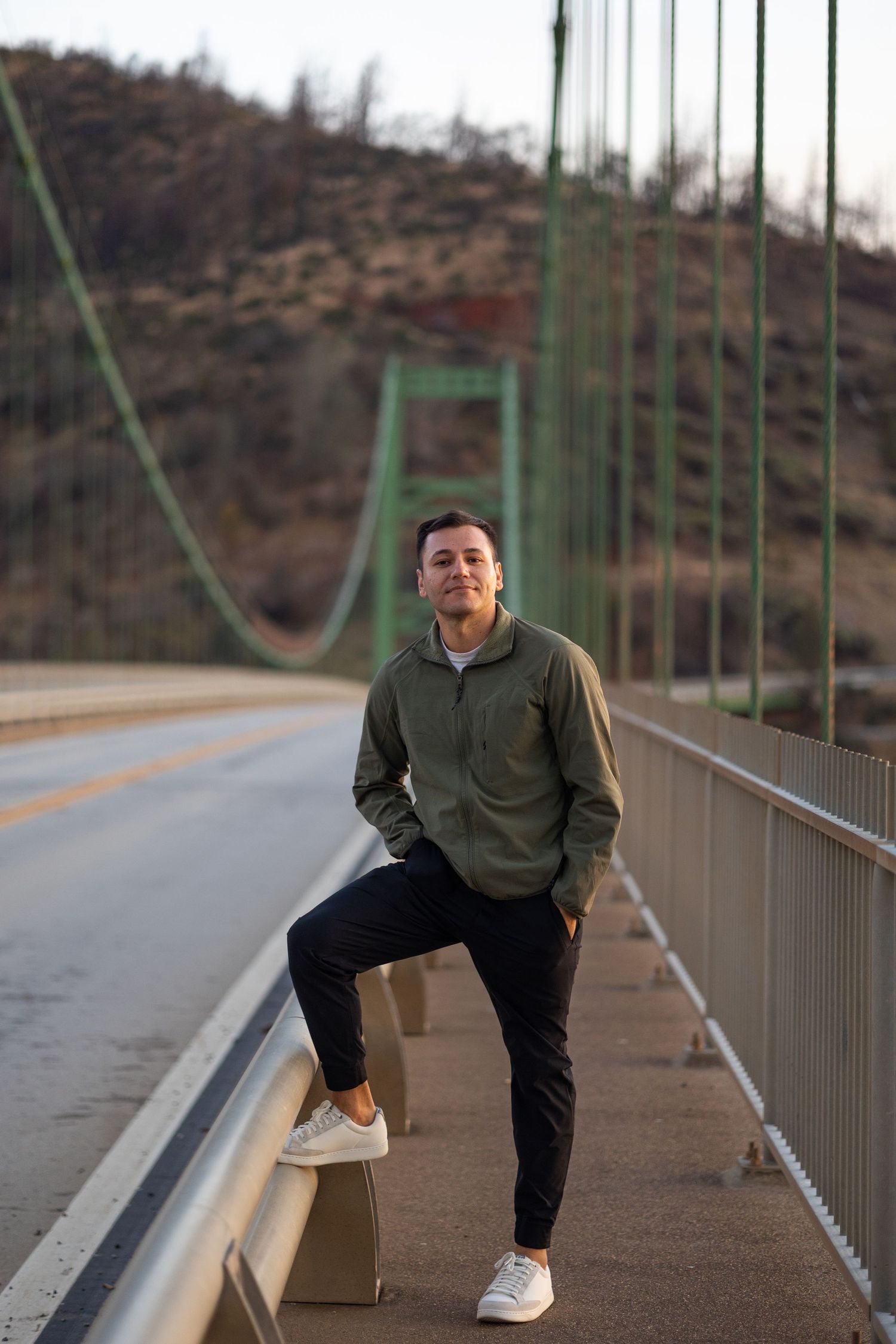 Young man in olive hoodie and black pants posing on a green suspension bridge.
