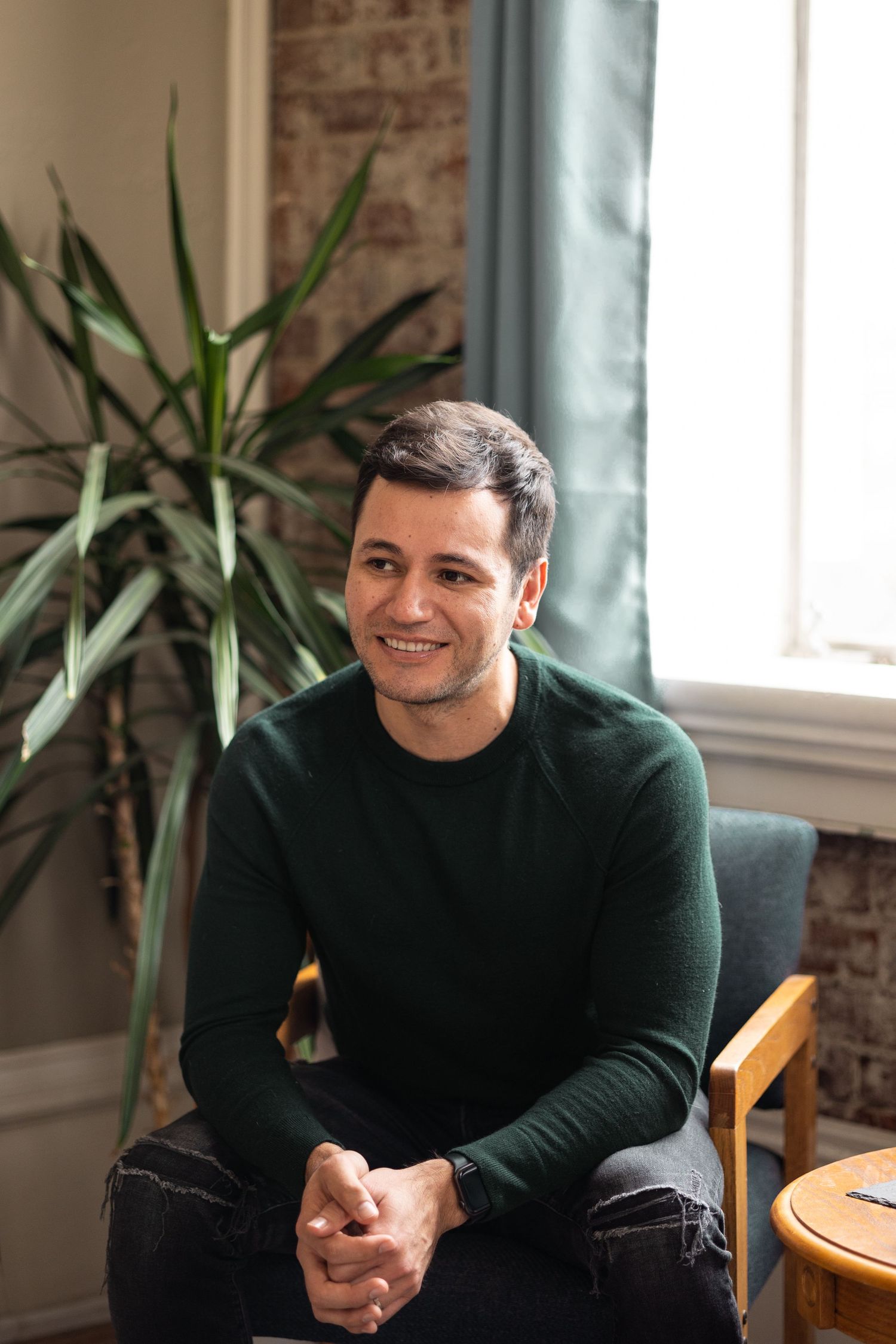 Smiling man in dark green sweater sitting in a chair near a window with indoor plant.