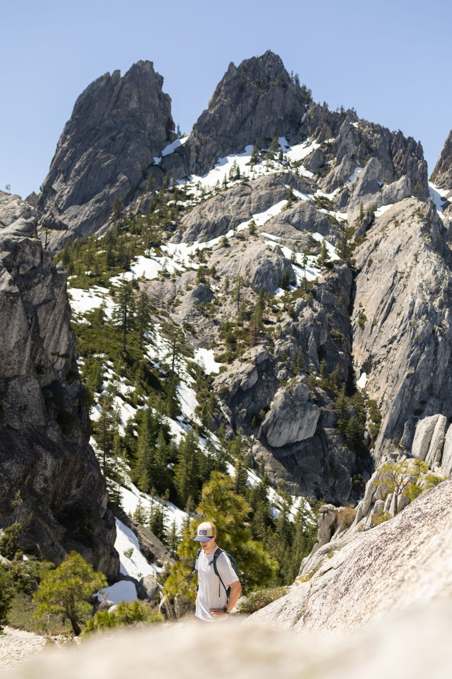 A hiker with a backpack walks along a rocky trail with dramatic snow-dusted mountain peaks behind.