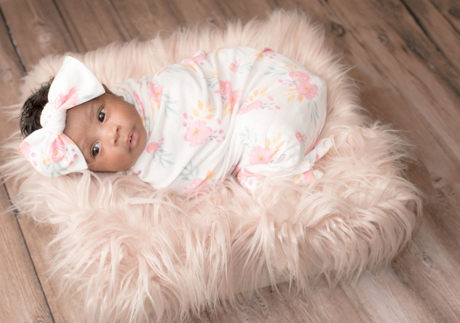 A newborn baby swaddled in floral print lying on pink furry backdrop.