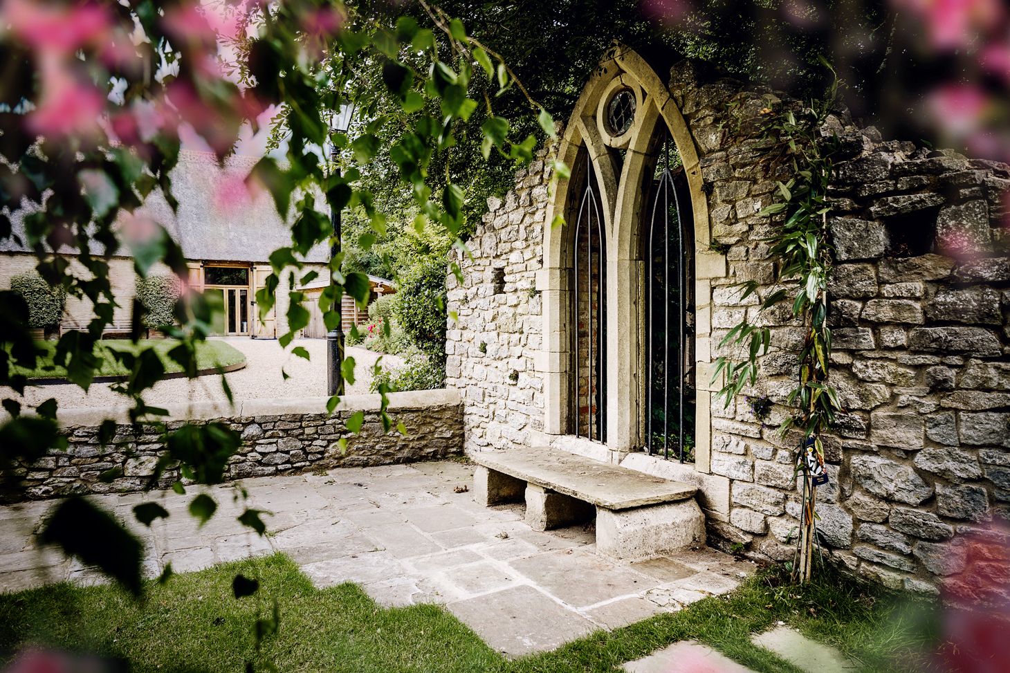 Ancient stone archway with Gothic window and bench in a garden setting framed by pink blossoms.