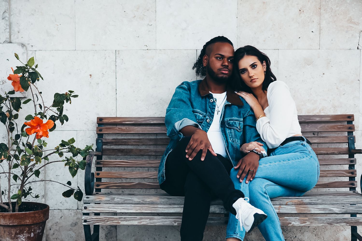 Young couple in denim jackets sitting together on bench near orange flowers.