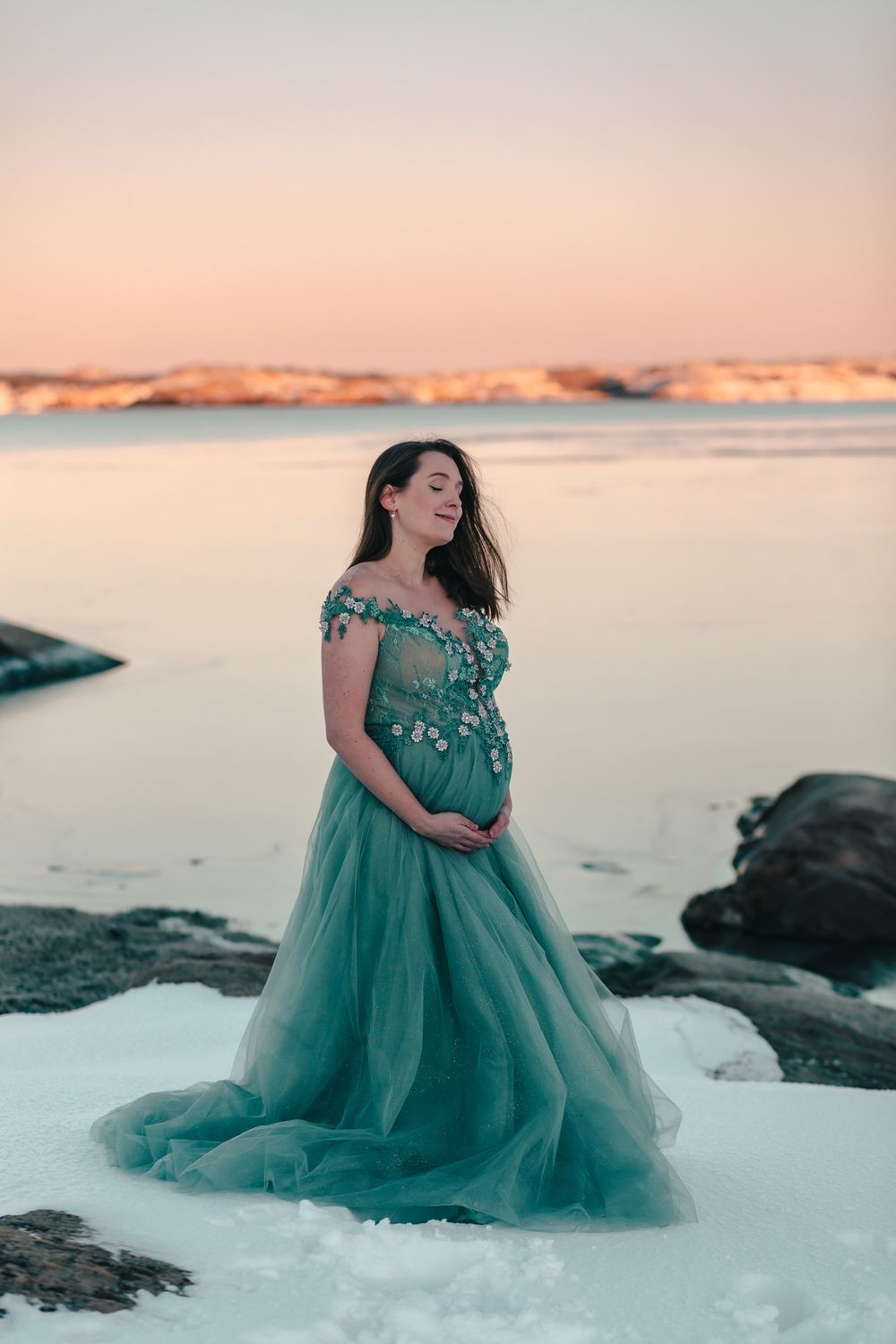 Person in flowing teal gown poses on snowy shore during sunset.