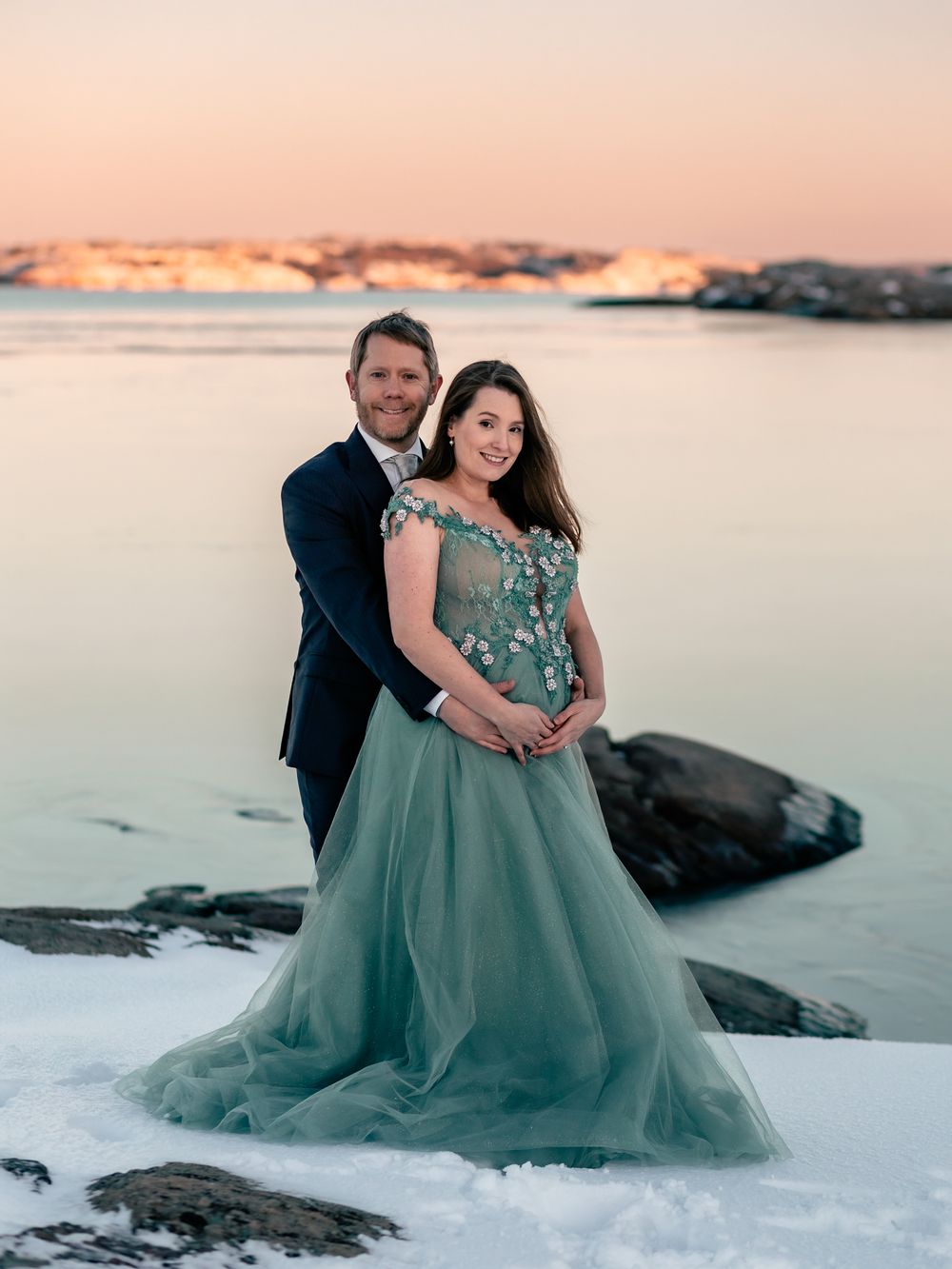 Couple in formal attire pose together on snowy beach at dusk.