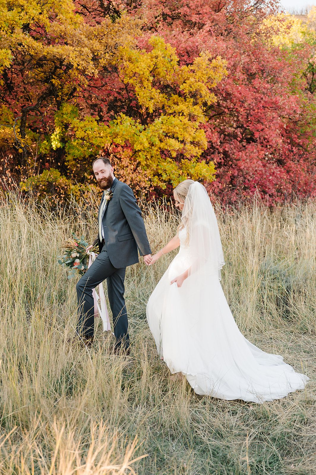 A wedding couple walks through tall autumn grass with vibrant yellow and red fall trees behind them.