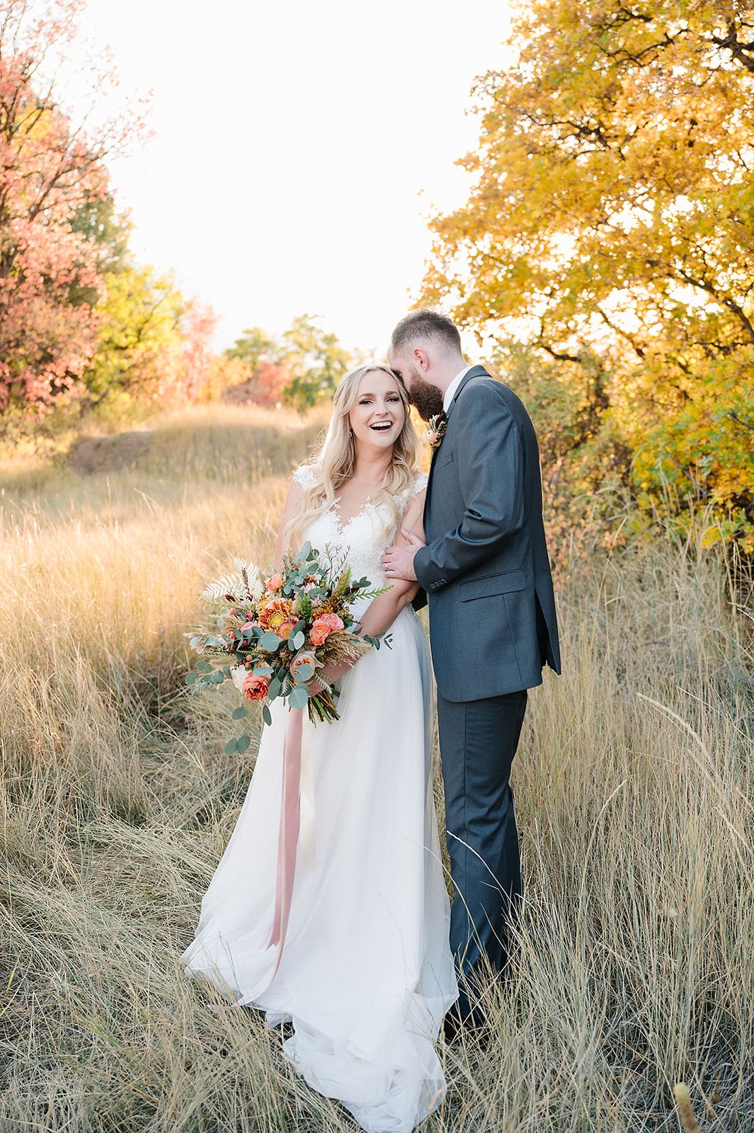A wedding couple shares a joyful moment in a golden autumn field with fall foliage.
