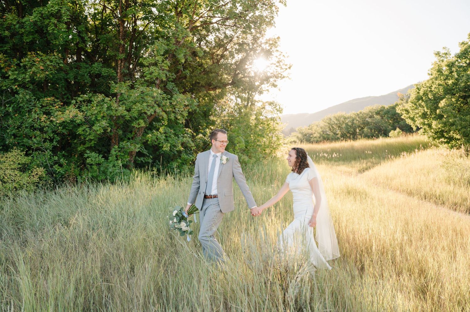 Couple holding hands while walking through golden meadow at sunset with mountains in background.