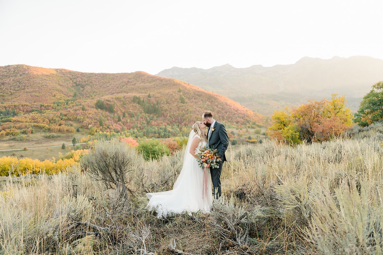 A wedding couple poses against a backdrop of mountains and desert brush at sunset.