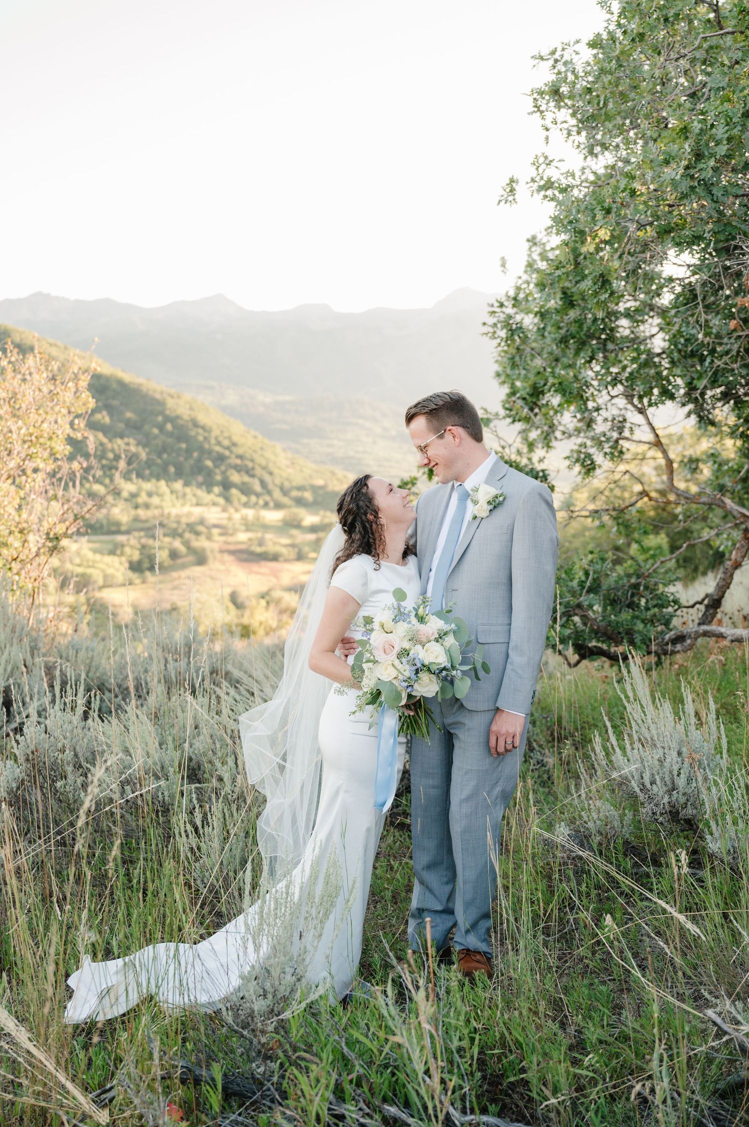 Couple in wedding attire embracing on mountainside with valley vista behind them.