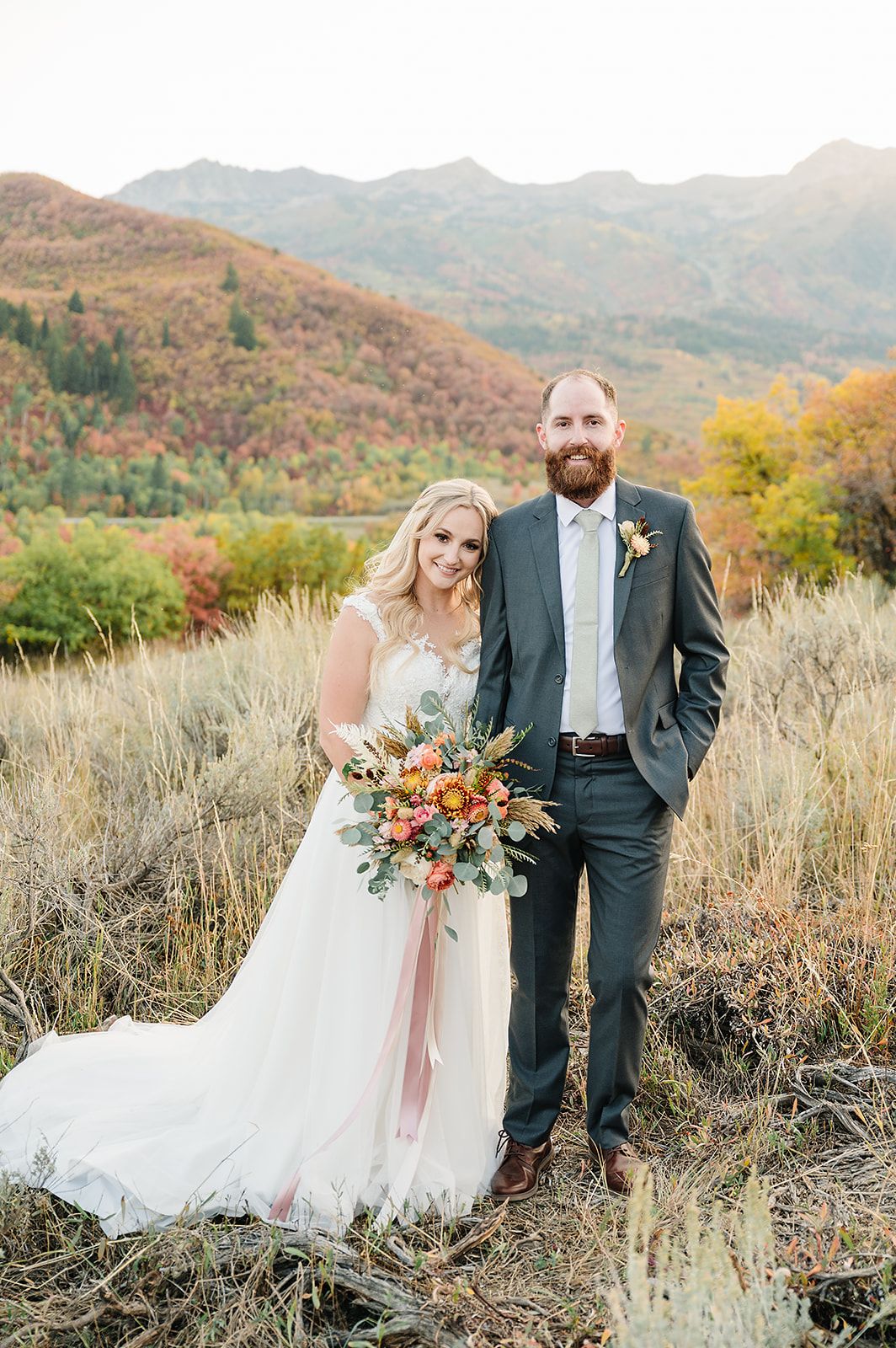 A wedding couple stands in a mountain meadow with autumn colors and rolling hills in the background.