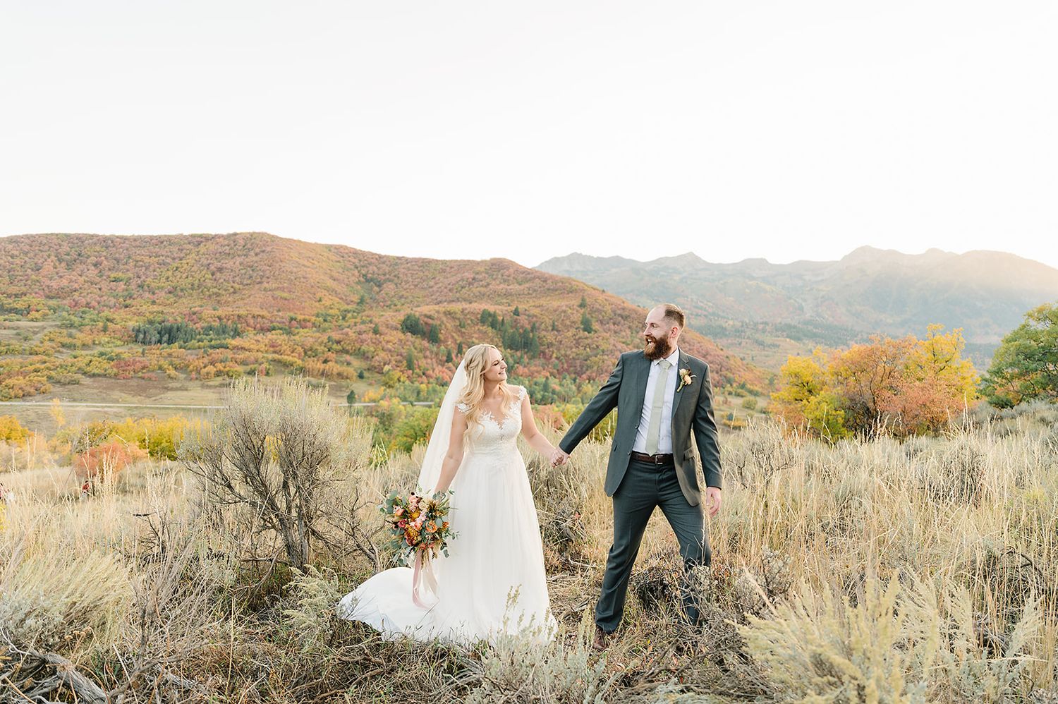 A wedding couple holds hands while walking through a mountain meadow at sunset.