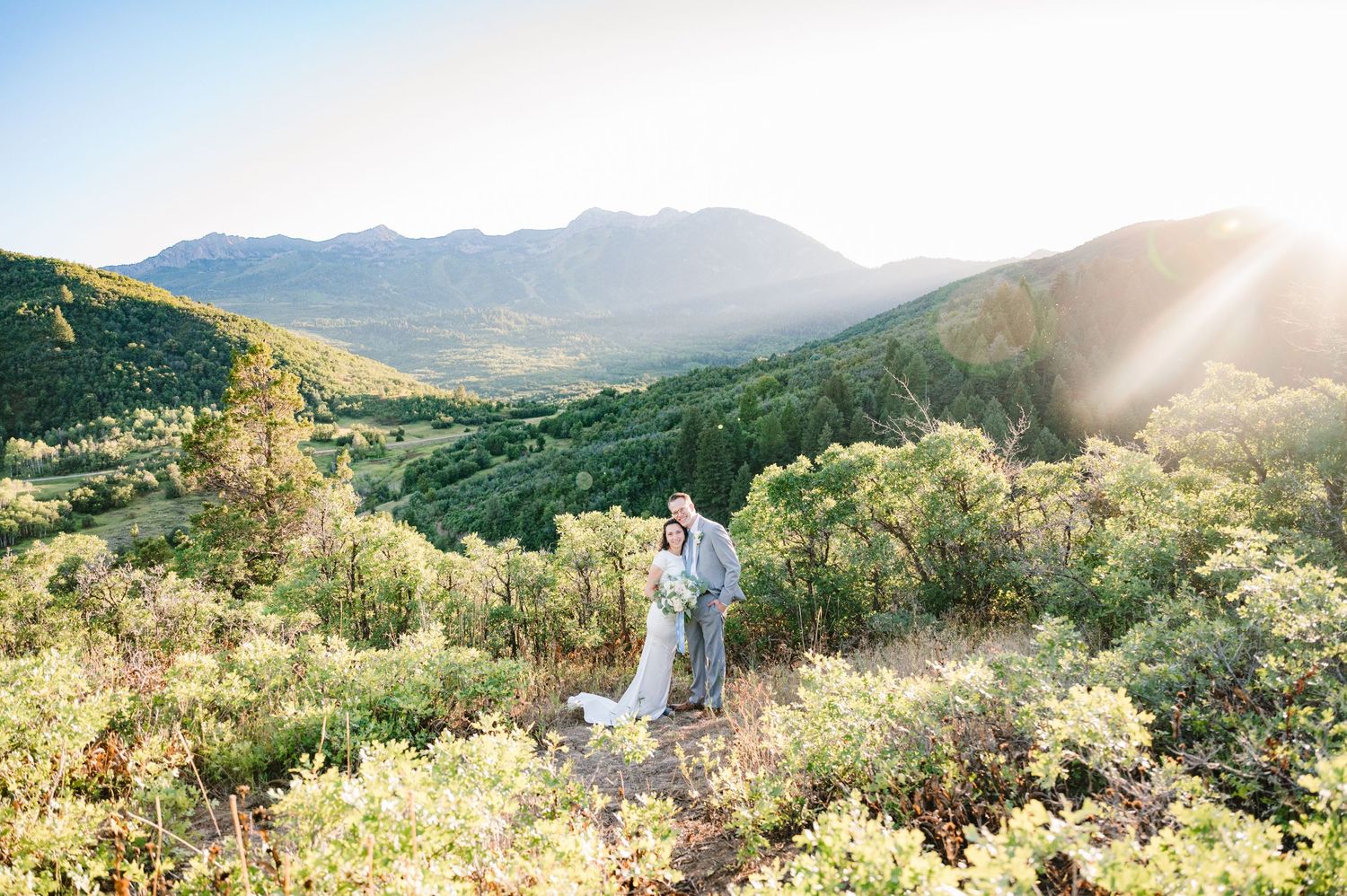 Wedding couple walking through mountain landscape at golden hour with dramatic sunburst.