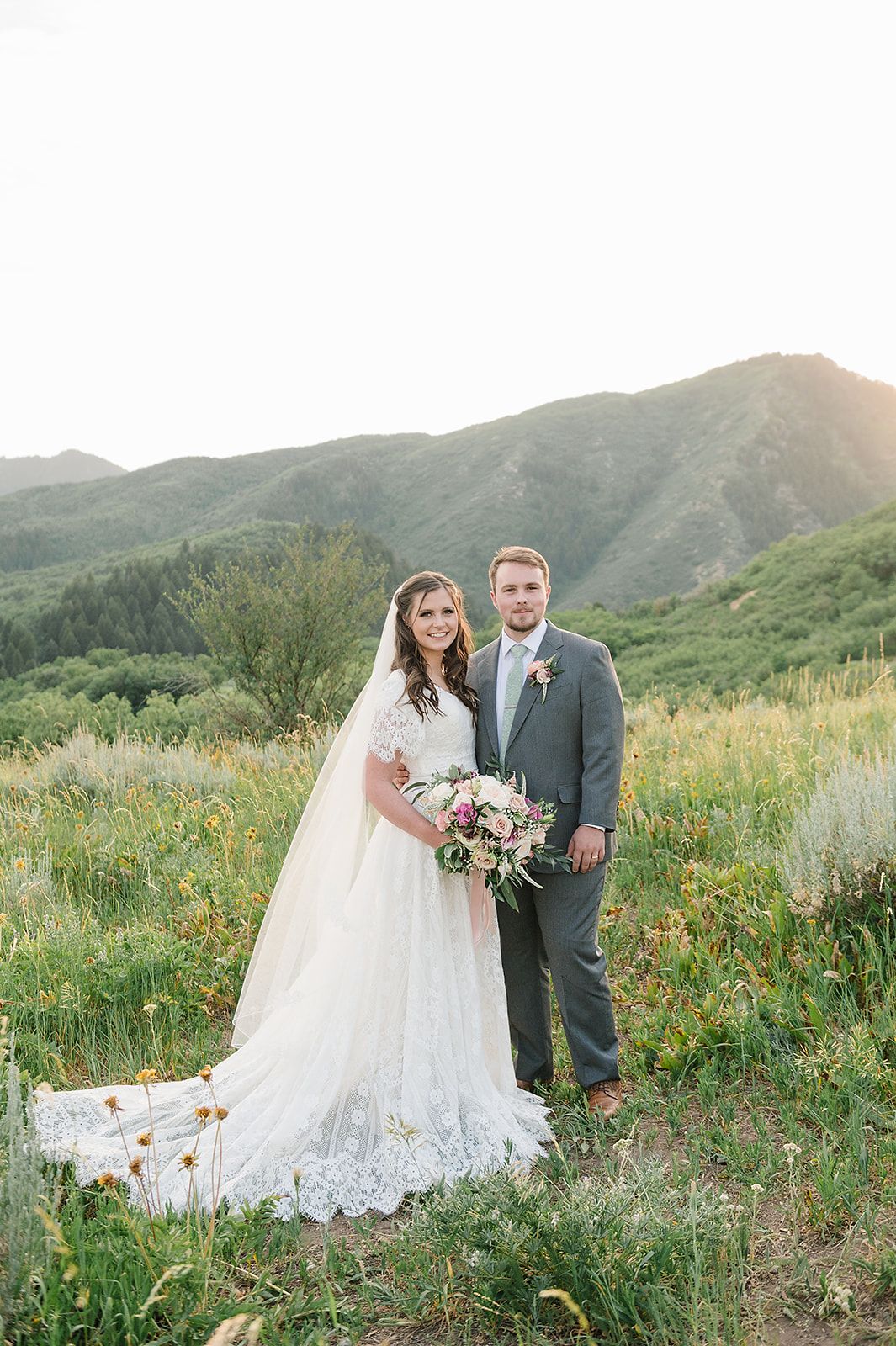 A wedding couple poses in a green mountain meadow with dramatic mountain peaks in the background.
