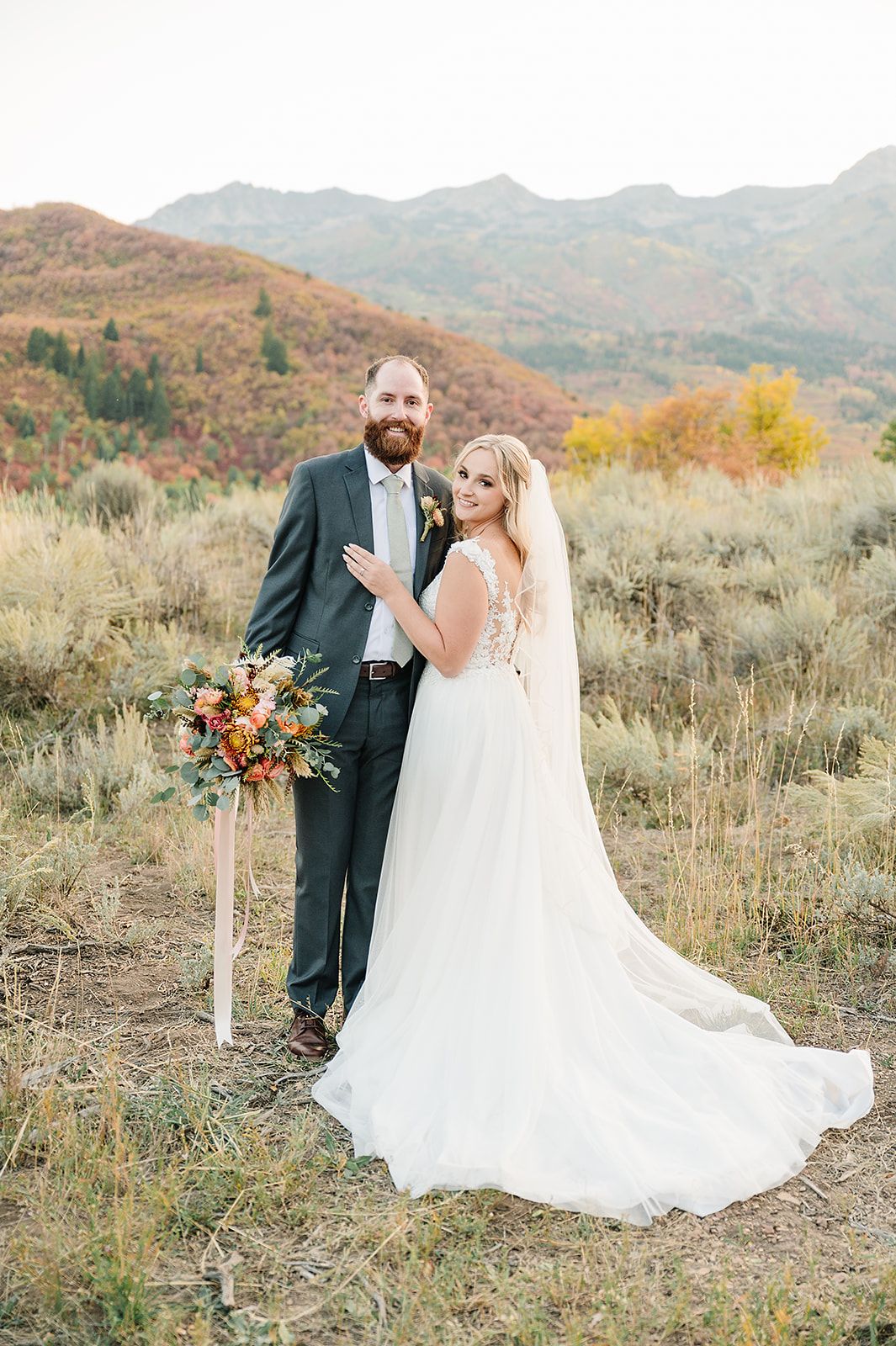 A wedding couple stands together on a mountain trail with autumn foliage and mountain views.