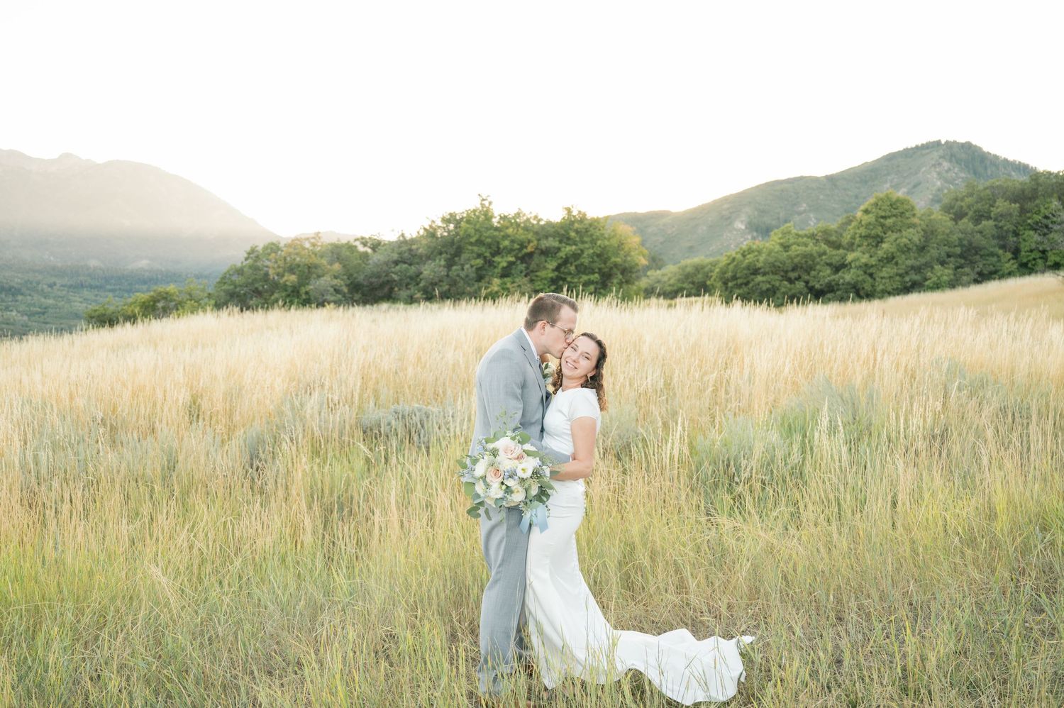Couple in wedding attire standing together in golden mountain meadow at sunset.