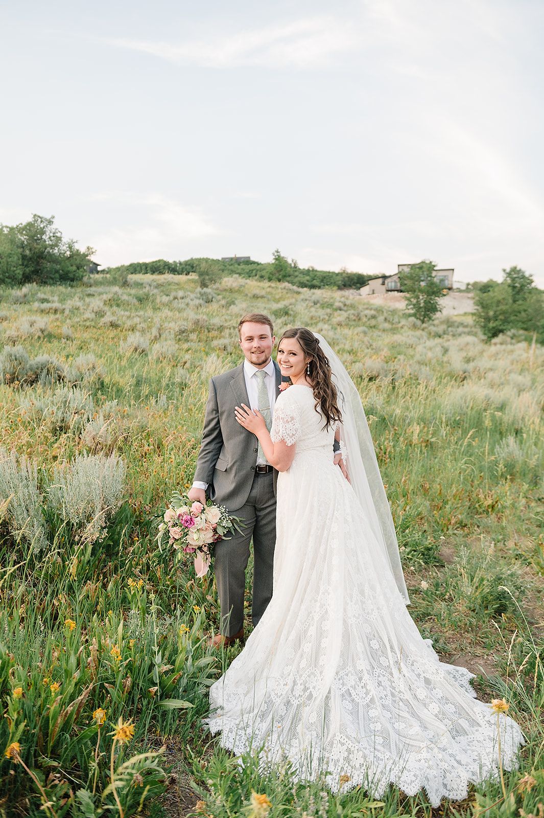 A wedding couple poses in a sunny meadow with wildflowers, the bride wearing a flowing lace gown with long train.