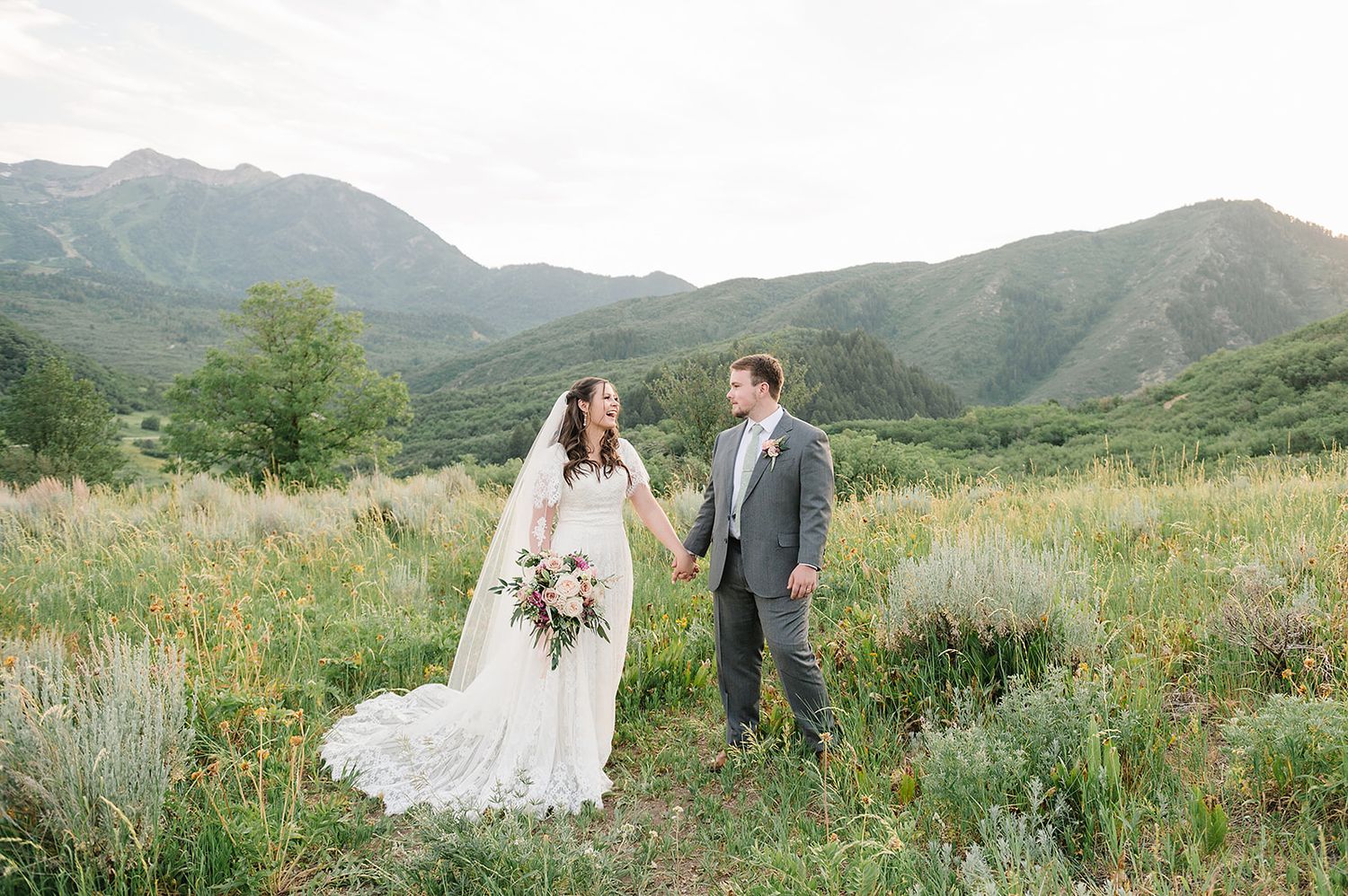 A couple in wedding attire stands in a mountain meadow with rolling hills and peaks in the background.
