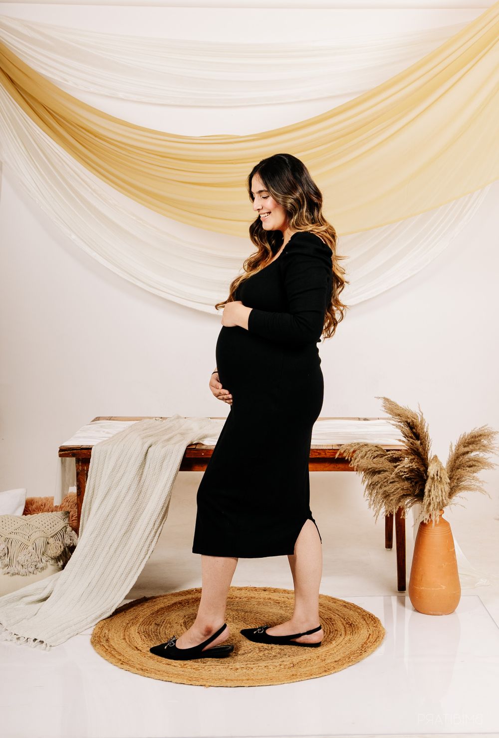 A maternity portrait with draped fabric backdrop showing a woman in black dress on a round jute rug.