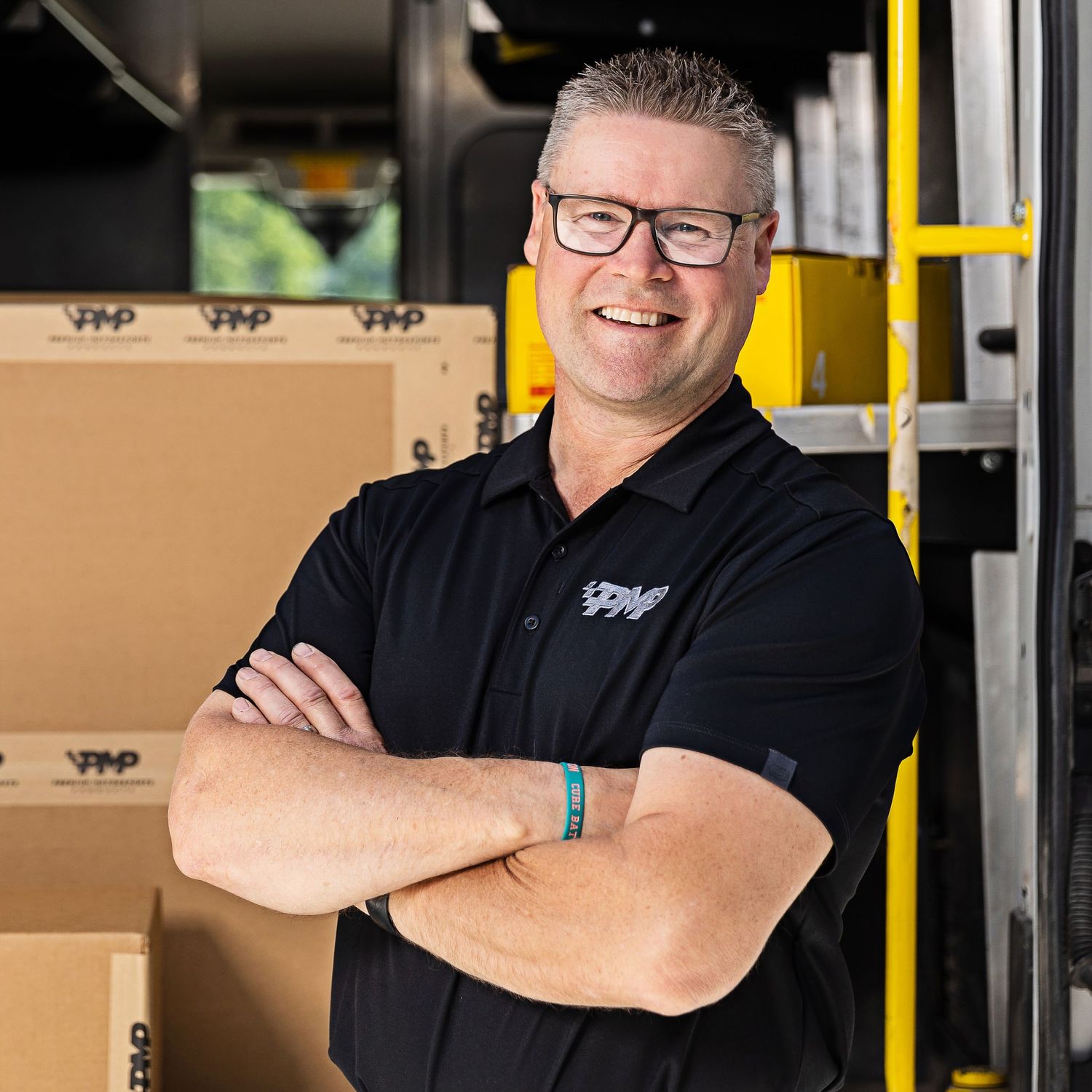 Warehouse worker in black polo shirt smiling with crossed arms near shipping boxes.