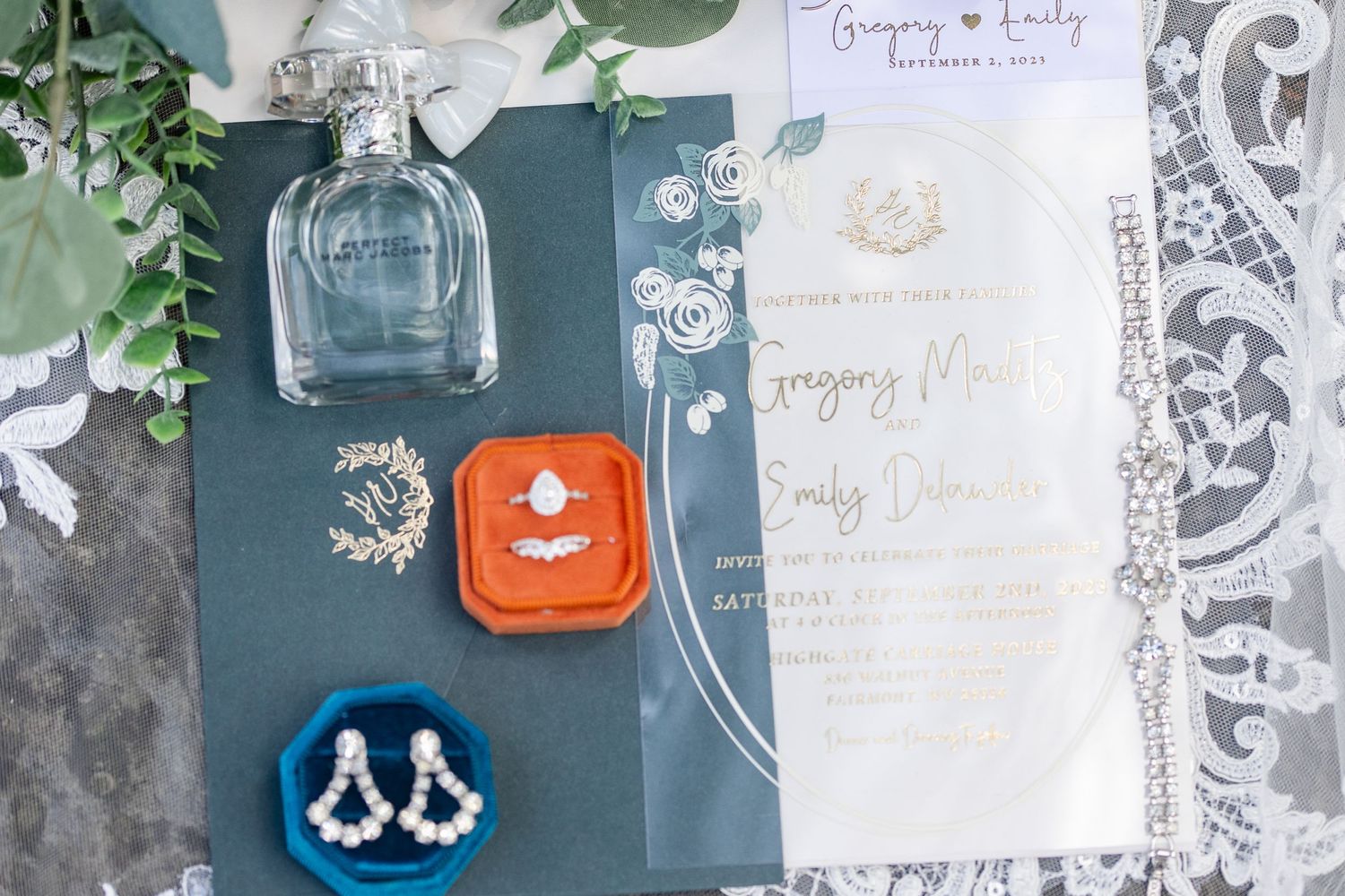 Wedding details flatlay with perfume bottle, jewelry, and invitation on dark background with eucalyptus.